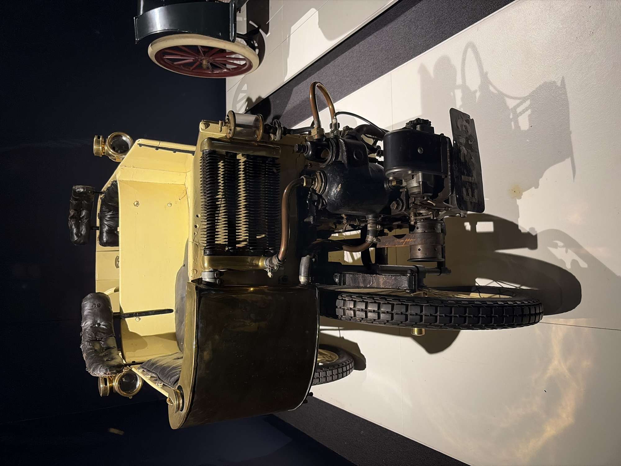 An antique, light yellow, open-engine motorized vehicle, likely an early automobile or quadricycle, is displayed indoors, presumably in a museum or exhibition. The photograph captures the vehicle from a slightly elevated side-angle, with the image rotated 90 degrees clockwise.
The vehicle features a prominent, exposed black engine block with numerous connecting copper pipes and tubing. A large, brass-colored finned radiator or cooling unit is positioned at the front. Below the engine, a large, dark metallic tank, possibly for fuel or oil, is visible. The vehicle has spoked wheels fitted with dark tires that have a distinct blocky tread pattern; one rear wheel is clearly visible on the right, and part of a front wheel is seen at the bottom. Brass-colored headlights or lanterns adorn the front corners of the body. Dark, padded sections, resembling leather upholstery, are visible along the sides, suggesting seating or armrests. A chain drive mechanism is clearly visible on the right side, connected to the rear wheel.
The setting consists of a light-colored floor and wall, contrasting with a dark background behind the vehicle. Shadows of the vehicle and its intricate components are cast on the bright surfaces, indicating artificial lighting designed for display. In the upper background, a portion of another antique vehicle is visible, specifically a wheel with red wooden spokes and a white-rimmed tire. No people or visible text are present in the image. The scene depicts a static exhibit highlighting early automotive engineering and design.