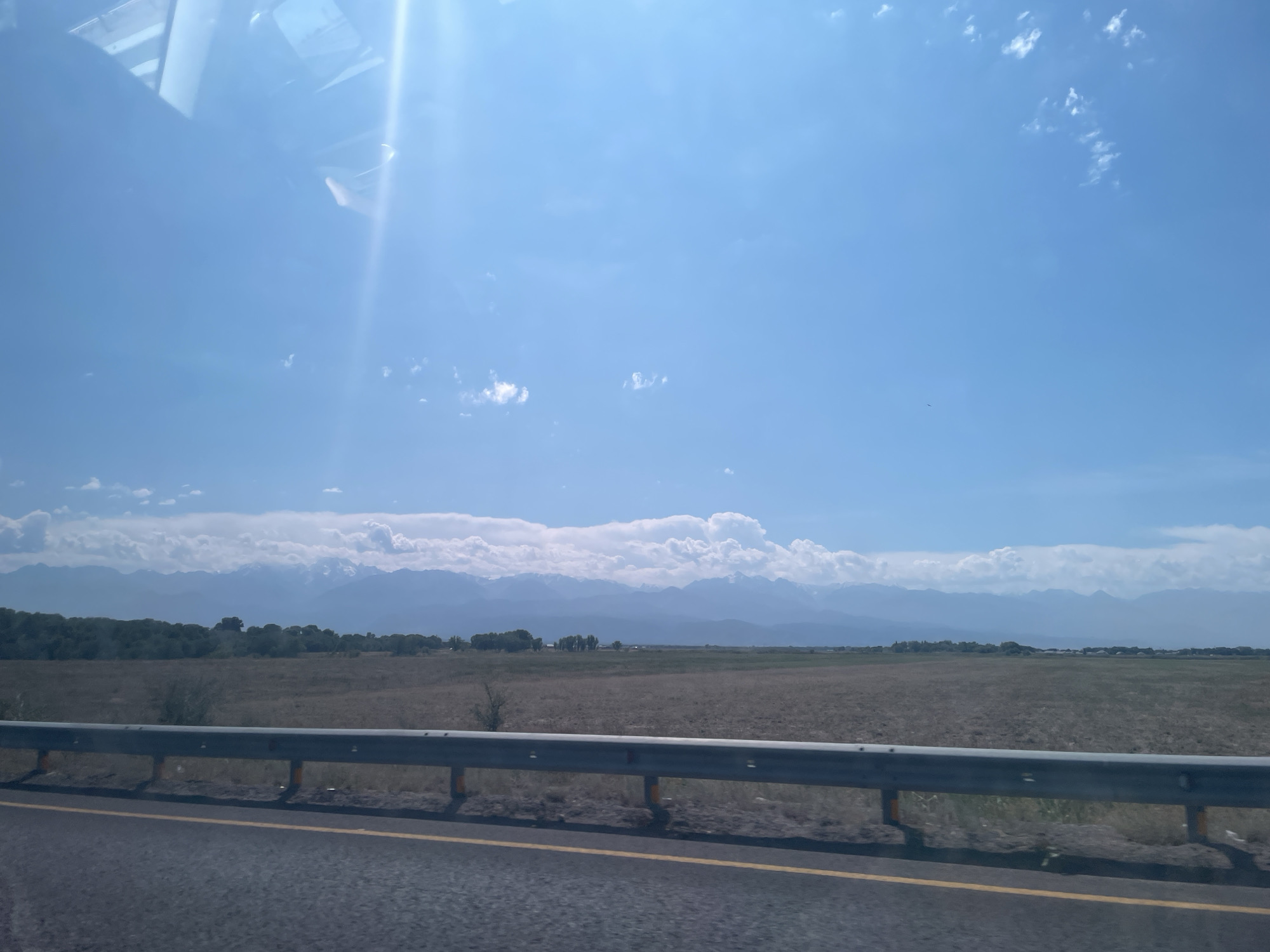 This image shows a view from a moving vehicle on a highway with a guardrail in the foreground. Beyond the guardrail is a wide, flat, dry field with scattered low bushes and trees. In the background, a range of majestic mountains capped with snow stretches across the horizon under a bright blue sky with scattered white clouds. The sun is shining intensely, creating a strong glare in the upper left portion of the image. The scene appears to be of the outskirts of Baiserke, Kazakhstan, likely captured during daylight hours under fair weather conditions. There are no people or specific actions depicted in the image.