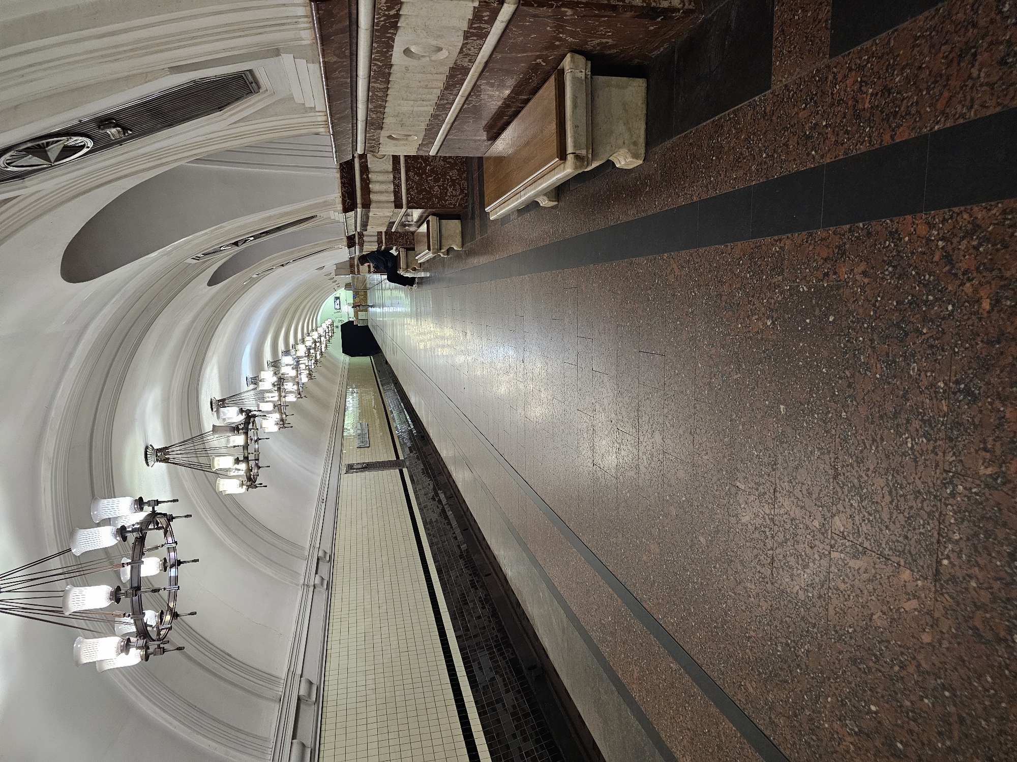 The image depicts a view of the Kiyevskaya metro station in Moscow, Russia. The station is notable for its ornate design, featuring a vaulted ceiling with multiple chandeliers and arches. The walls are covered in white tiles, and the floor is a mix of marble and granite. Benches line one side of the platform. At least one person is visible, seated on one of the benches.