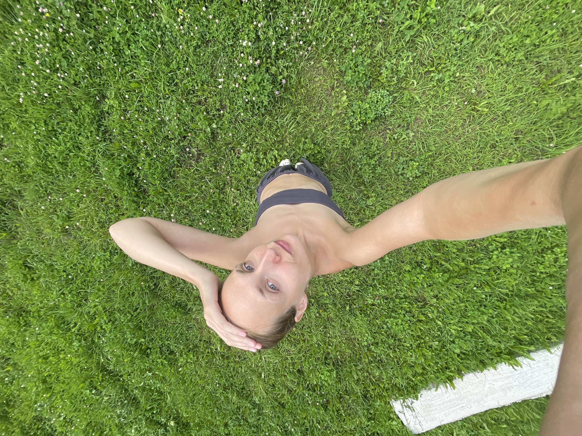 A person is lying on a grassy area with small white flowers scattered throughout. They are taking a selfie from a high angle, looking up at the camera with their head tilted back and hands on their head. The person is wearing a dark sports bra and black shorts. Their arms are outstretched to the sides. The scene appears to be outdoors on a bright day, as indicated by the lighting and green grass. The overall mood is relaxed and casual. There is a white rectangular object partially visible in the lower right corner of the frame, which could be a towel or mat.