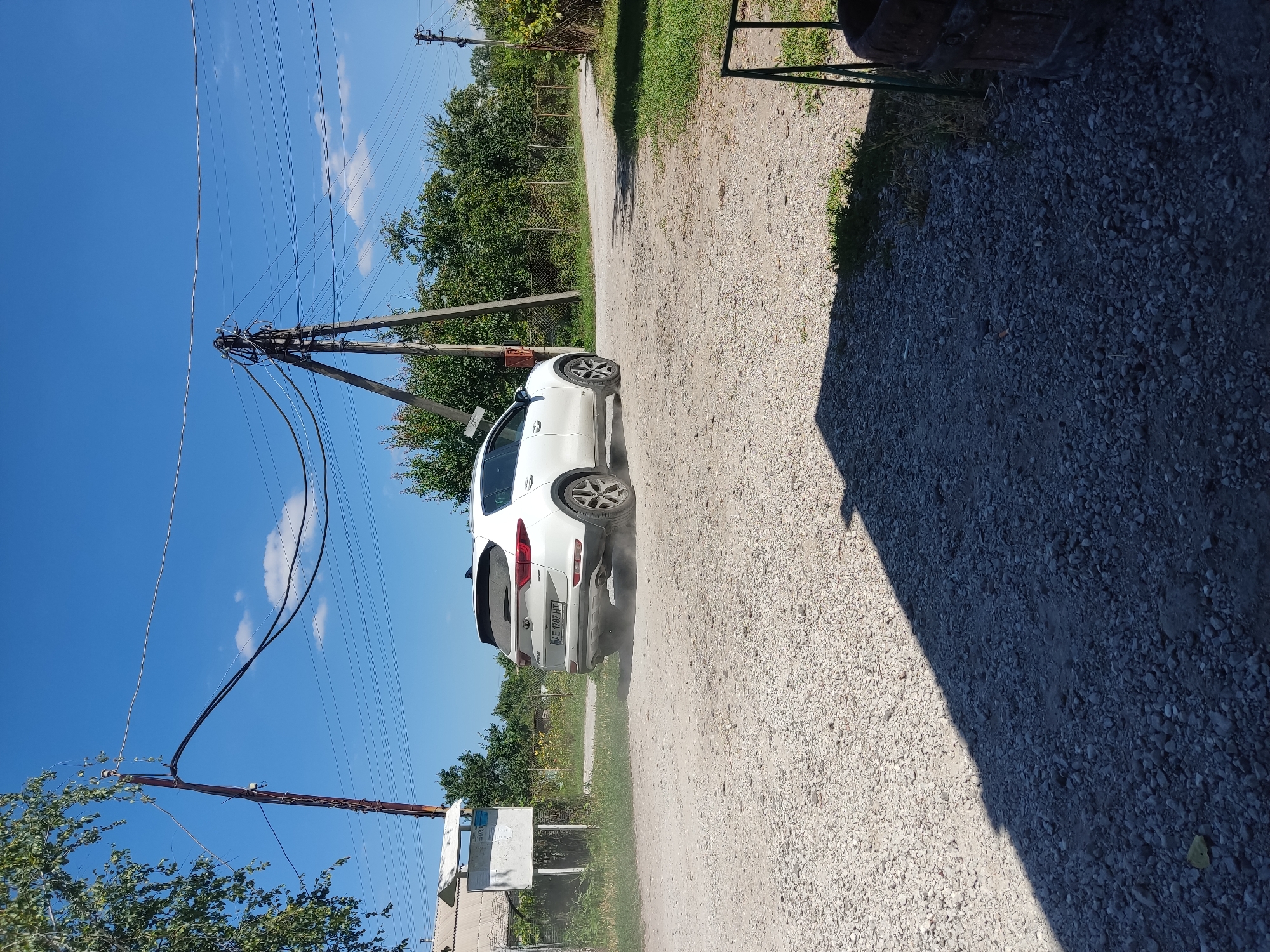 A white Kia Sportage is seen from the rear-right driving away on a gravel road, kicking up dust. The vehicle has a license plate that reads "AE 1787 HT". The scene is outdoors under a clear blue sky with some scattered white clouds. In the background, there are trees, power lines supported by wooden poles, a fence, and a small building with a signpost. A shadow of a structure falls across the foreground. The setting appears to be a rural or semi-rural area, likely in the village of Radisne, Ukraine, given the context.