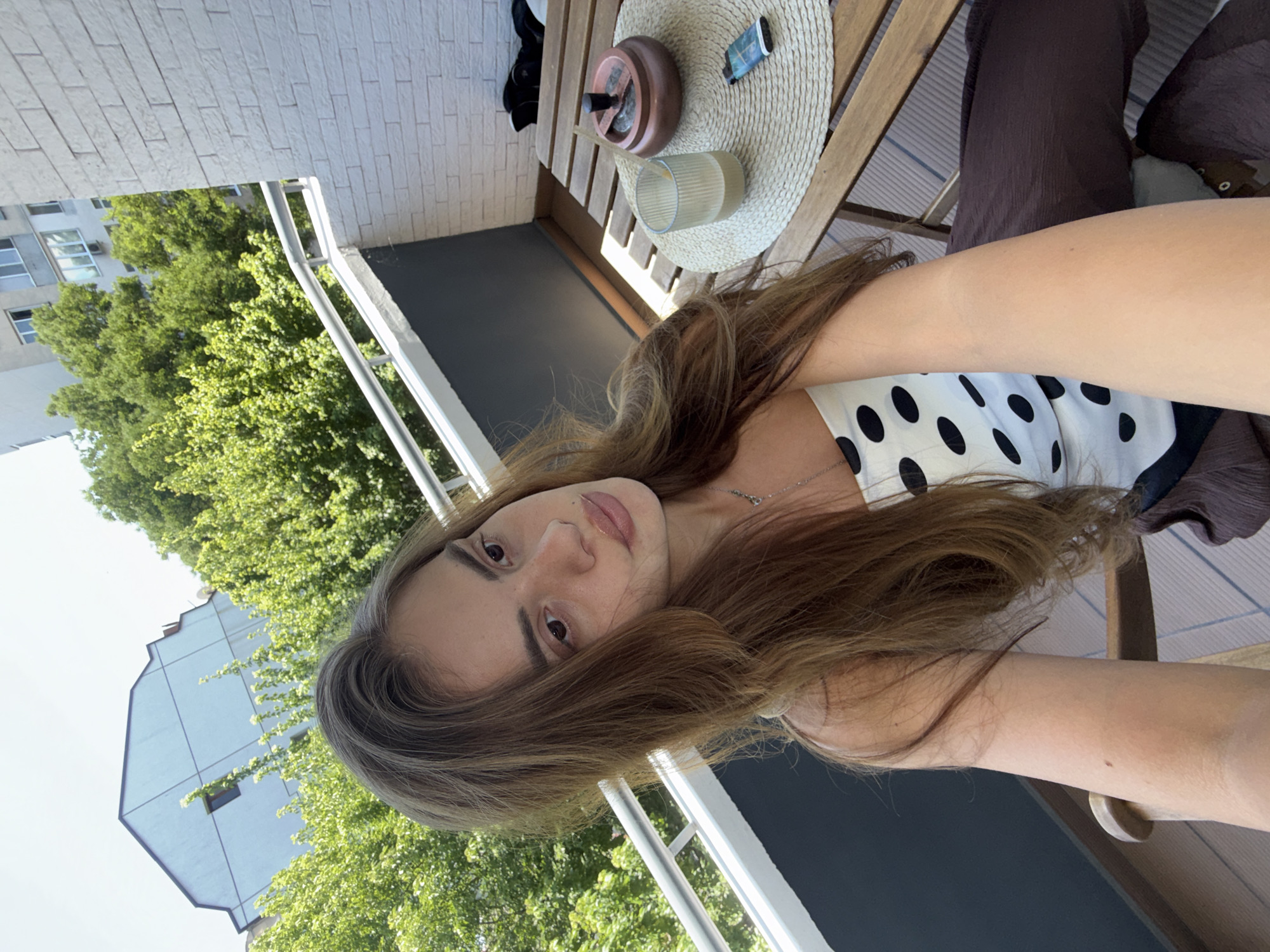 The media file shows a young woman with long, brown hair taking a selfie on a balcony. She is wearing a white top with black polka dots. The setting is a balcony with a wooden table and chairs. On the table, there is an ashtray, a glass, and a lighter. Behind her, there are trees and a building. The photo appears to be taken during the day, with bright sunlight. The woman has a relaxed expression. The context provided indicates the location is Plovdiv, Bulgaria.