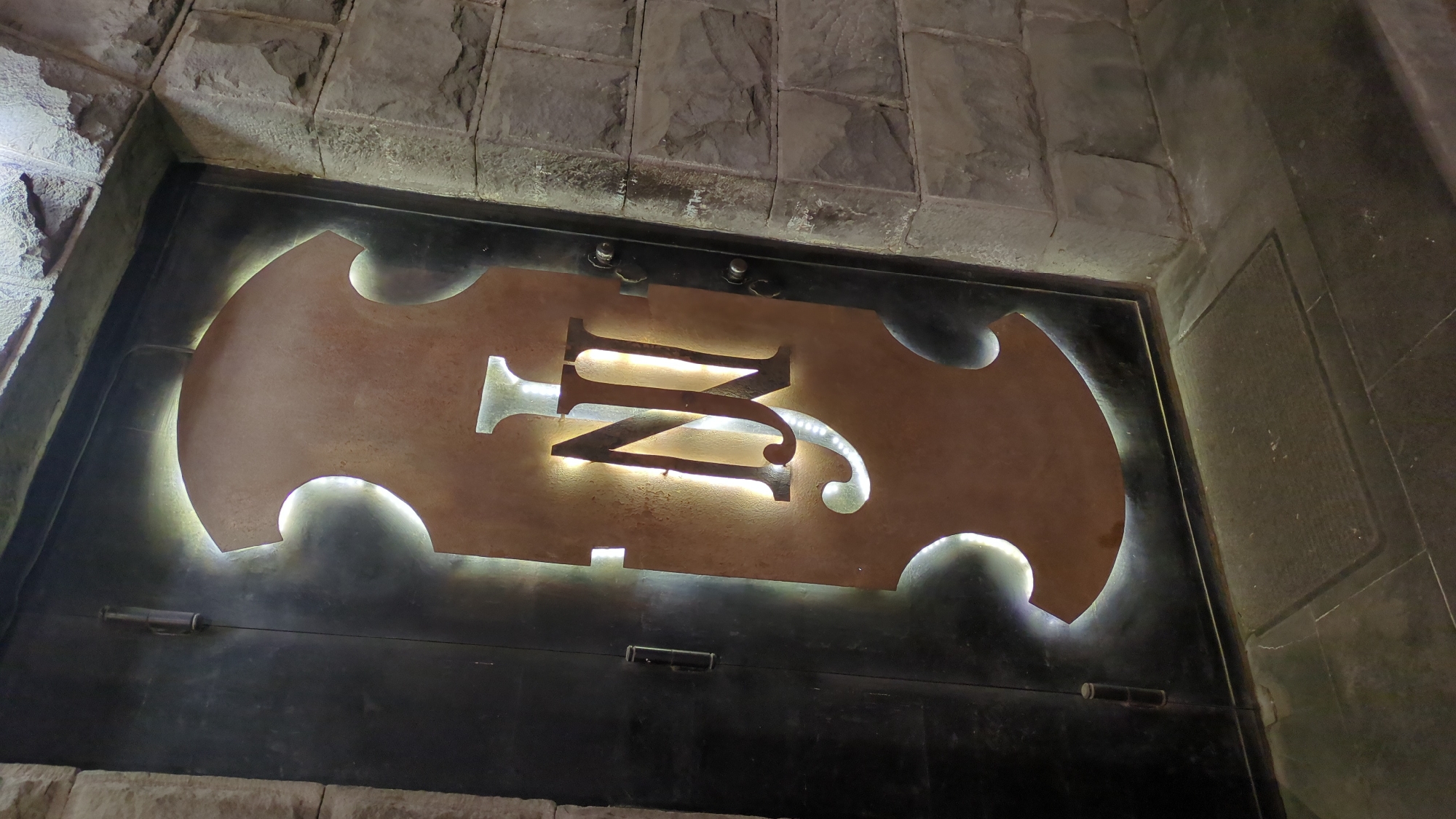 The image displays a prominent, backlit metal sign featuring an intertwined "M" and "N" monogram, set against a dark background, possibly a door or a large panel. The sign's main body is made of a rustic, bronze-toned metal with an ornate, curved outline, resembling a decorative shield or the body of a musical instrument. Bright white light emanates from behind the cut-out letters, creating a striking halo effect that illuminates the edges of the monogram and the textured surface of the sign.
The dark panel behind the sign, which appears to be dark metal, exhibits visible hinges along its lower edge and mounting fixtures at the top, suggesting it is a substantial door. This door or panel is framed by a robust architectural structure composed of large, rough-hewn gray-brown stone blocks, indicative of an archway or an entrance. The contrast between the natural stone and the custom metalwork is notable.
The scene is dimly lit, suggesting an indoor environment, evening, or night, where the illuminated sign acts as a primary source of light and a focal point. There are no people visible, and no specific activity is taking place; the image focuses on the static architectural detail. This distinctive custom signage is located in Yerevan, Armenia, adding to the character of the establishment it marks.