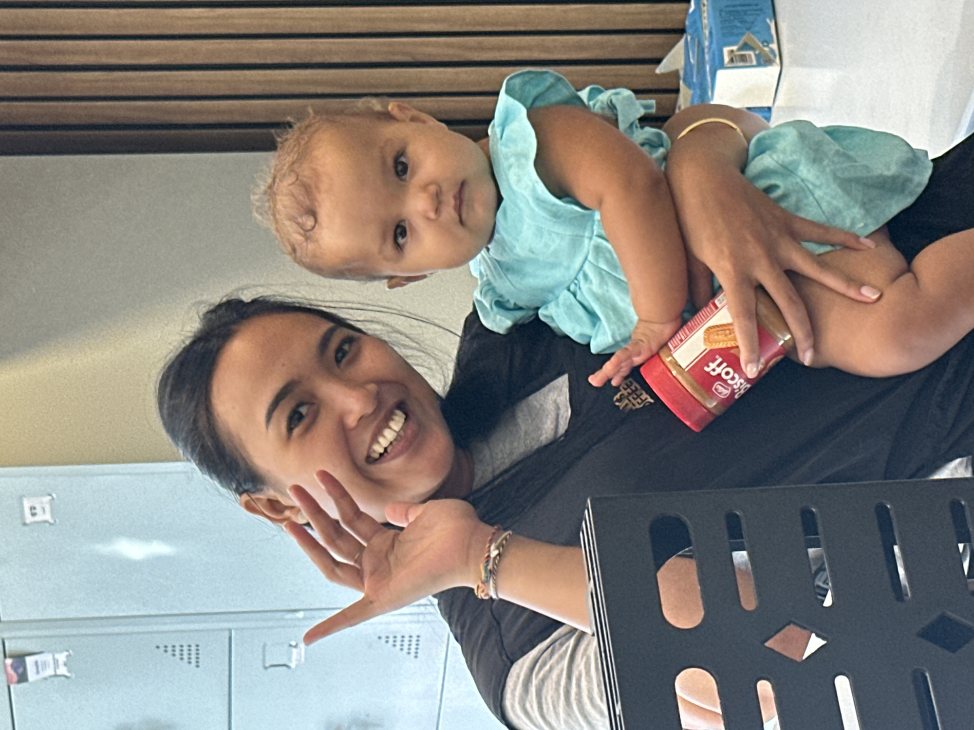 A medium shot captures a woman holding a baby in a room with lockers in the background, possibly a school or office. The woman, with dark hair pulled back and a bright smile, is wearing a grey t-shirt and a black vest. She is looking at the camera and waving with her left hand, which is adorned with several bracelets. The baby, wearing a light blue ruffled dress, is being held in the woman's arms and looking forward. The woman is holding a jar of Biscoff spread in her right hand, partially obscured by the baby. The environment features light blue lockers with white markings and a wooden paneled wall in the background, suggesting an indoor setting. The overall impression is a candid moment of affection between the two.