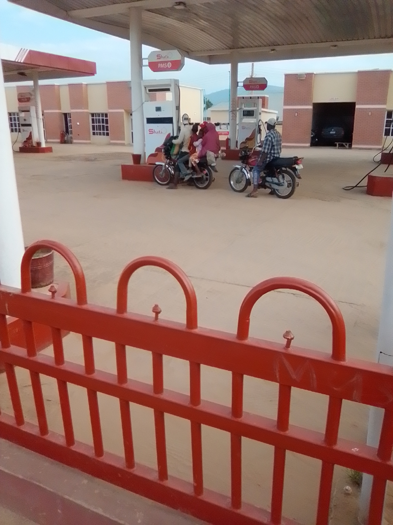 The image depicts a street view of a Shafa filling station in Kwadon, Nigeria, under a bright, overcast sky. The station features red and cream-colored buildings with numerous windows, and large overhangs supported by white pillars. Several fuel pumps are visible, with one white pump prominently displaying the Shafa logo and "PMS" label.
In the foreground, a red metal fence with decorative arches and pointed finials obstructs the view of the street. Beyond the fence, three motorcycles are parked. One motorcycle, a red model, is occupied by a man wearing a plaid shirt and a white cap, facing away from the camera. Another motorcycle, also red, is ridden by two individuals, one in a light-colored top and the other in a dark top with a red hood. A third motorcycle, a silver model, is also present.
The ground is paved with a light-colored material, possibly concrete or asphalt. In the background, a dark-colored car is parked inside a building structure, suggesting a service bay or garage. The overall scene conveys a sense of everyday activity at a local transportation hub.