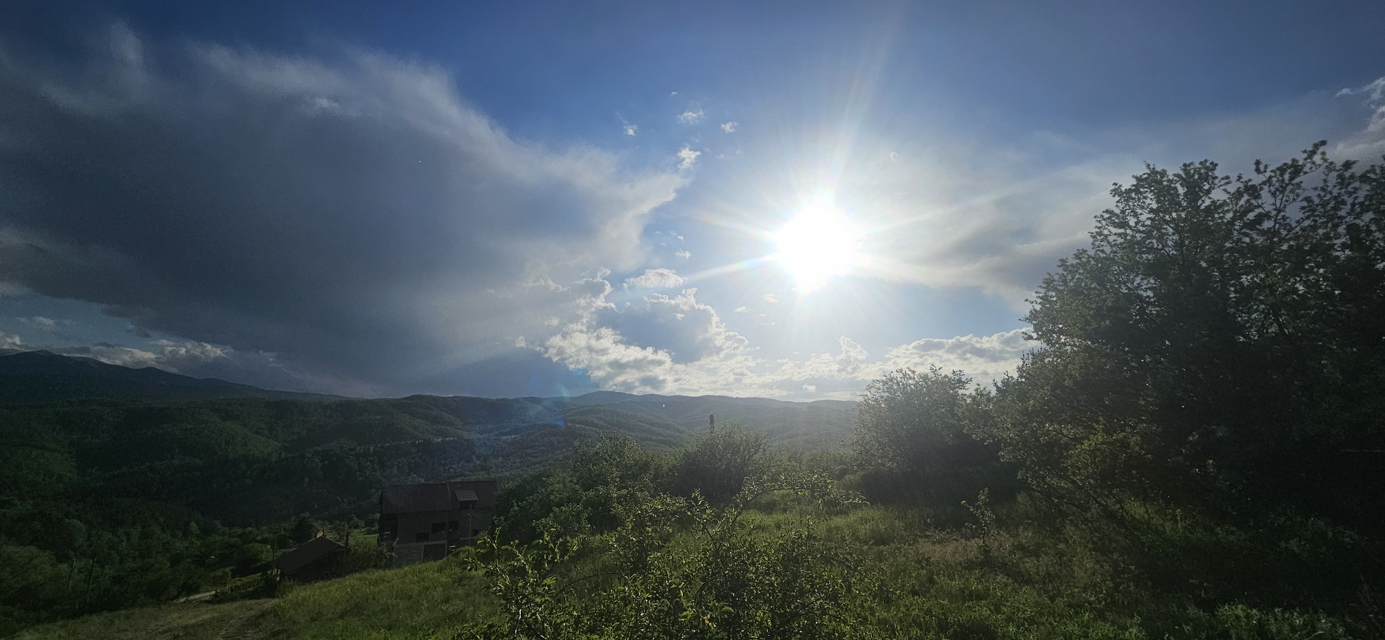 The image depicts a scenic landscape in Gutsal, Bulgaria. The sky is a mix of blue and heavy, dark storm clouds. The sun is high and bright, shining through the clouds. Below, a lush green hillside is covered in vegetation and trees. A few houses are visible nestled in the hillside. In the distance, mountains extend along the horizon.