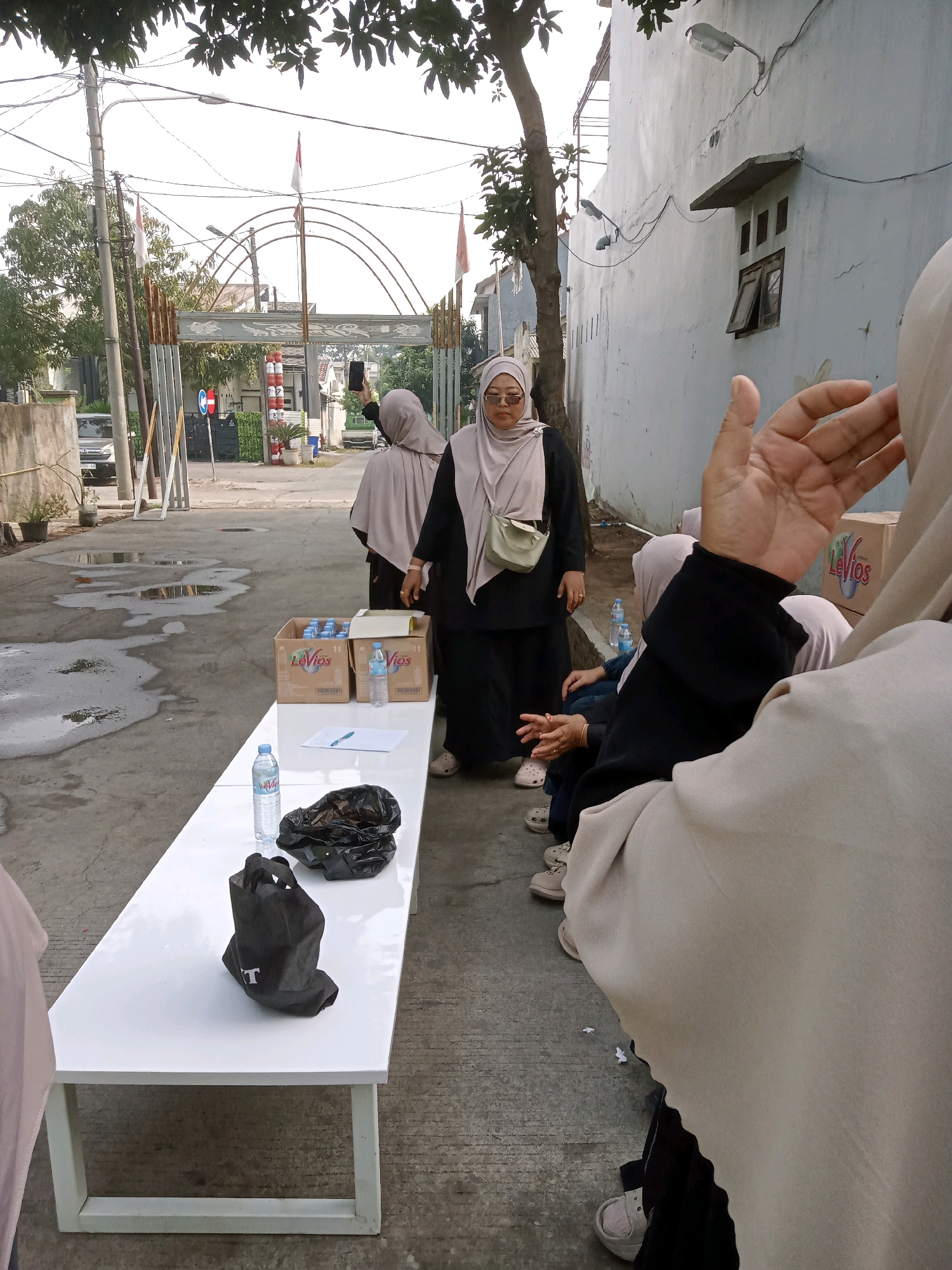 The image depicts an outdoor scene in a residential or commercial alleyway in Karawang, Indonesia. The foreground is dominated by a long, white table on which a bottle of water, a black plastic bag, and a black shopping bag with a logo are placed. Several people, mostly women wearing hijabs and modest clothing, are visible. In the background, there is an arched gateway with Indonesian flags, suggesting a possible event or entrance to a complex. Boxes of bottled water are stacked on the ground behind the table, and a few cars are parked in the distance. The ground is paved, with some puddles of water indicating recent rain or a damp environment. A building with a light-colored wall and a window is on the right side of the image. The overall atmosphere appears to be casual, with some individuals observing or interacting with the scene.