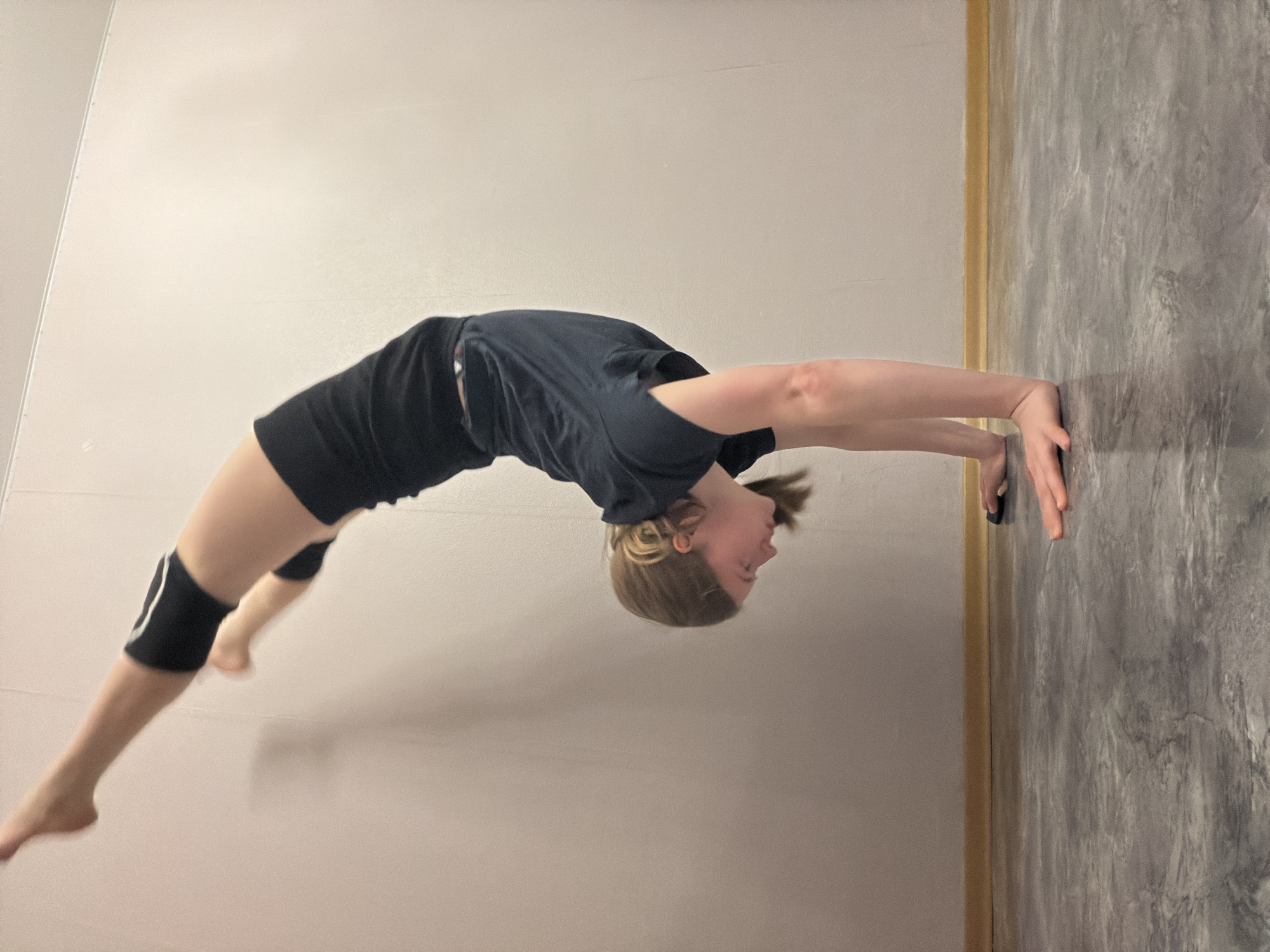 A girl in a dark t-shirt and shorts, with knee pads on her left leg, is performing a gymnastic backbend with her hands touching a gray wall. She is in a room with a white wall to her left and a gray textured wall to her right. The location is unknown, and there is no evidence to indicate it is in Perm, Russia.