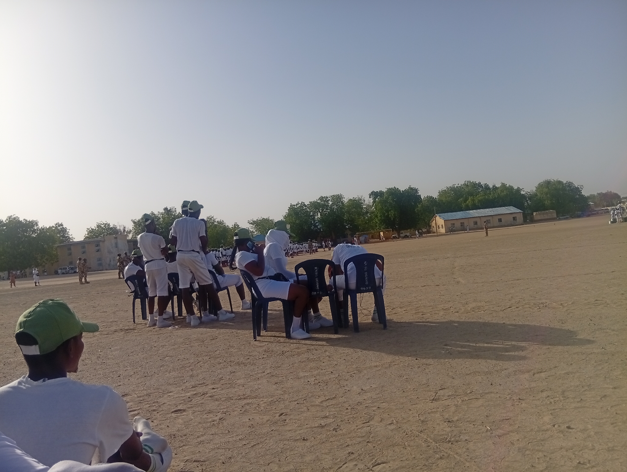 The image depicts a group of people in a dirt field. Most are dressed in white shirts, white shorts, and white socks and shoes, and some are wearing green baseball caps. A few people are sitting in black plastic chairs. There are a few buildings and trees in the background. The location is likely in Maiduguri, Nigeria, suggested by the text "NYSC BORNO" printed on the back of the chairs. The people appear to be participants in a formal event.