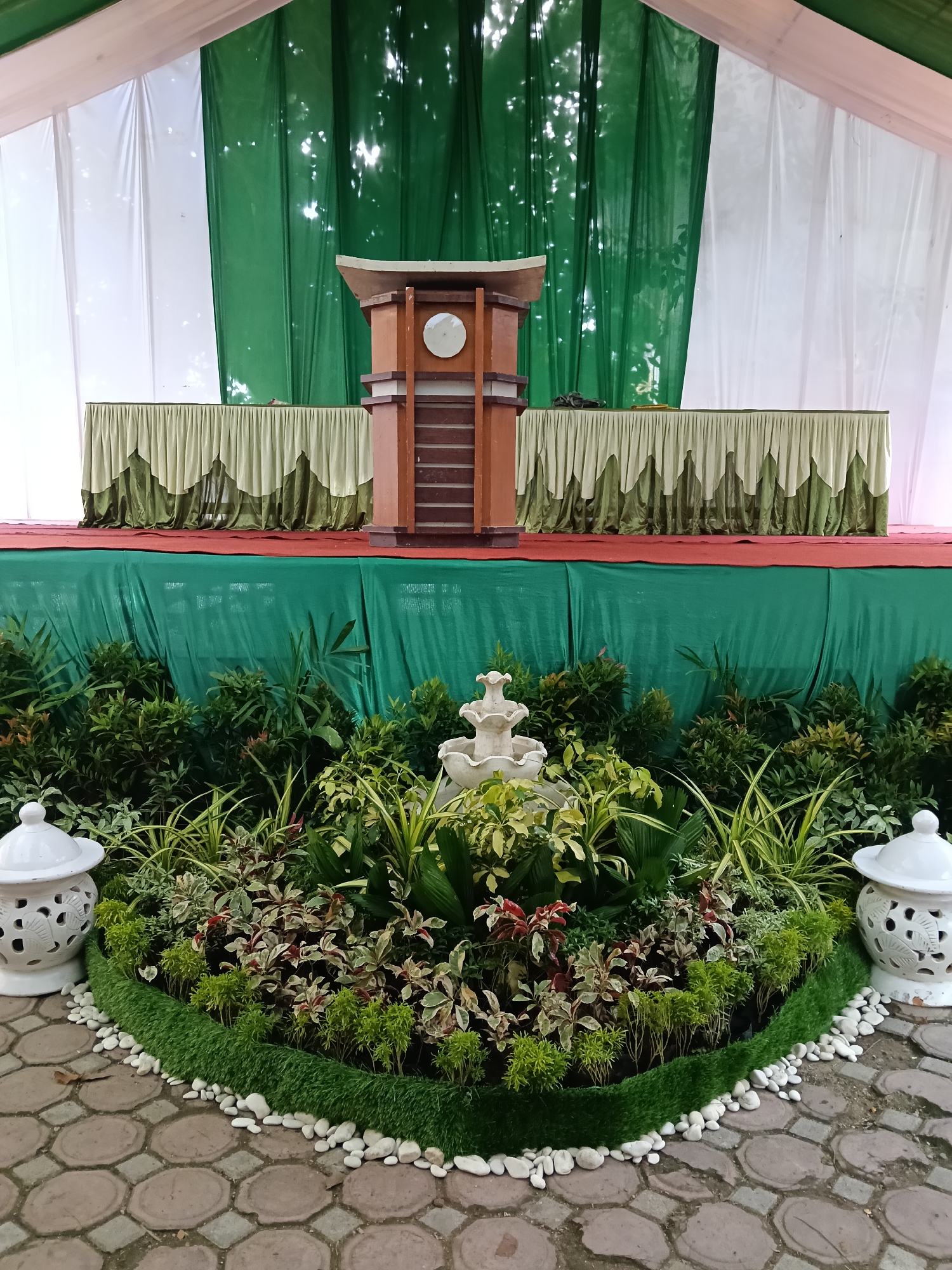 This media file captures a carefully arranged stage and foreground setup, likely prepared for an event. The central feature is a wooden podium with a curved top and a circular light-colored emblem on its front, situated on a stage covered with a red carpet. Behind the podium, a long table is elegantly draped with a light green skirt featuring a decorative scalloped border in a darker shade of green. The stage's backdrop consists of large drapes, alternating between white and deep green, with sunlight reflecting off the green fabric, indicating a daytime setting, possibly under a tent.
In front of the stage, a semi-circular garden bed is meticulously designed with a variety of lush plants, including various shades of green, variegated foliage, and some plants with reddish hues. A small, white, tiered decorative fountain is nestled among the plants, adding a serene element to the arrangement. The edge of the garden bed is outlined with a strip of artificial green turf and bordered by numerous smooth white pebbles, which contrast with the paved ground. The ground itself is constructed from octagonal interlocking bricks. Two ornate white ceramic lanterns with intricate cut-out patterns stand on either side of the plant bed, complementing the decorative scheme.
No people are visible in the image, but the elaborate and formal arrangement of the stage, podium, and decorative elements strongly suggests preparation for a formal or semi-formal event, such as a speech, presentation, or ceremony. The overall aesthetic conveys a sense of order and welcome. There is no visible text within the image. Given the location context of Lhokseumawe, Indonesia, the green and white color scheme and traditional podium align with common event aesthetics in the region.