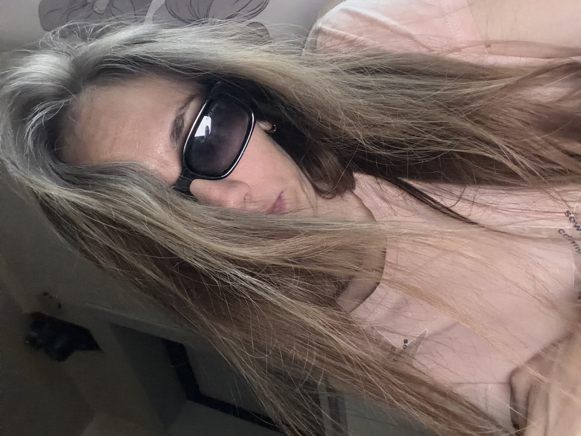 A woman wearing sunglasses and a pink t-shirt poses for a selfie indoors. The background is blurred, but hints of a room with a patterned curtain and what appears to be a shelf with items can be seen. The lighting is dim, casting shadows and highlighting the texture of the woman's blonde hair, which partially obscures her face. The image is a close-up, focusing on the woman's upper body and face. There are no visible indicators of the city of Tambov, Russia.