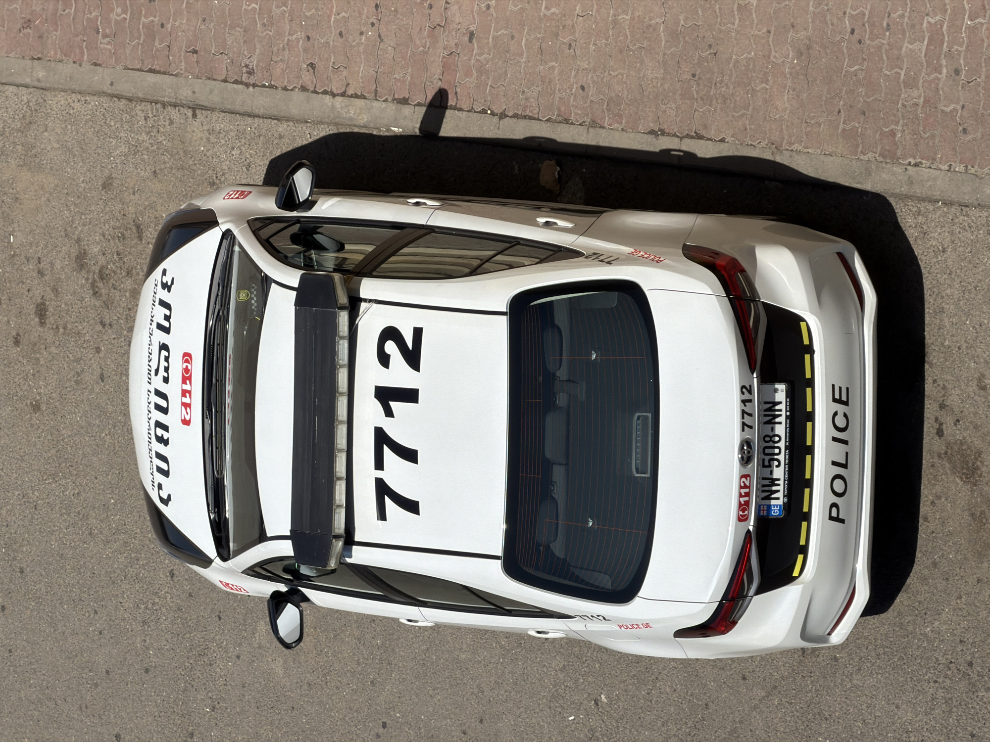 Here is a forensic description of the scene:
The image shows an aerial view of a white Georgian police car parked on a paved surface, likely a street or parking area. The license plate reads "NW-508-NN". Markings on the car indicate it belongs to the police ("POLICE") and contains the number "7712" prominently displayed on the roof. Additional smaller markings include "112" and "POLICE.GE". The car features black stripes on the roof and rear. Georgian text is visible on the front of the car. The car is casting a dark shadow on its right side. The image appears to be taken outdoors during daylight hours. The text on the car includes the address of Toyota Center Tegeta at University Street in Tbilisi.