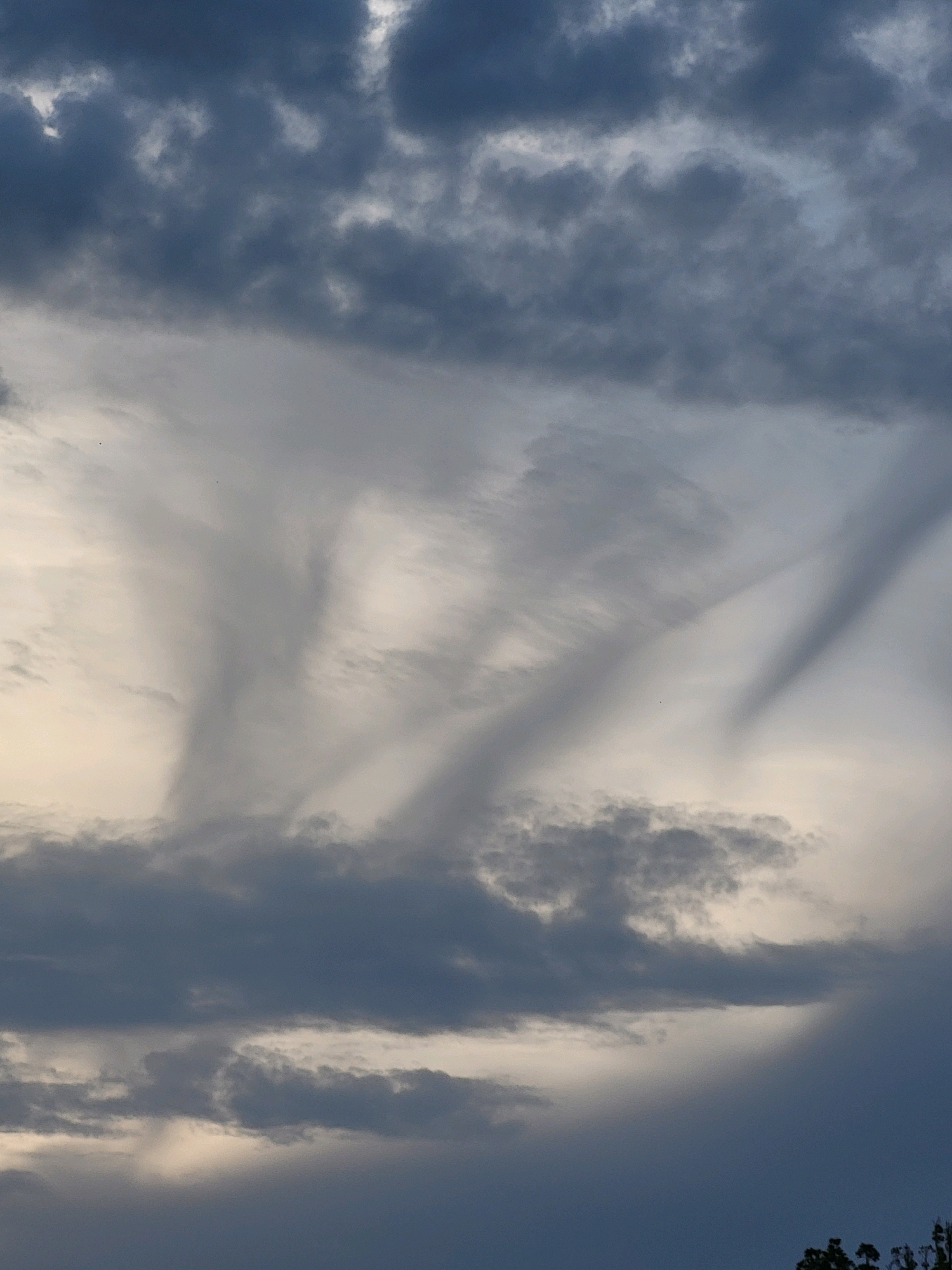 The image shows a sky with a variety of cloud formations. Dark, heavy clouds are present in the upper and lower portions of the frame, with lighter, wispy clouds forming streaks that fan out across the middle. The lighting suggests it may be near dawn or dusk. The scene appears to be an outdoor view, with the possibility of trees visible at the very bottom edge of the frame, suggesting a natural or semi-natural environment. There are no people, animals, or man-made structures clearly identifiable within the frame. The image was taken in Ozolnieki, Latvia.