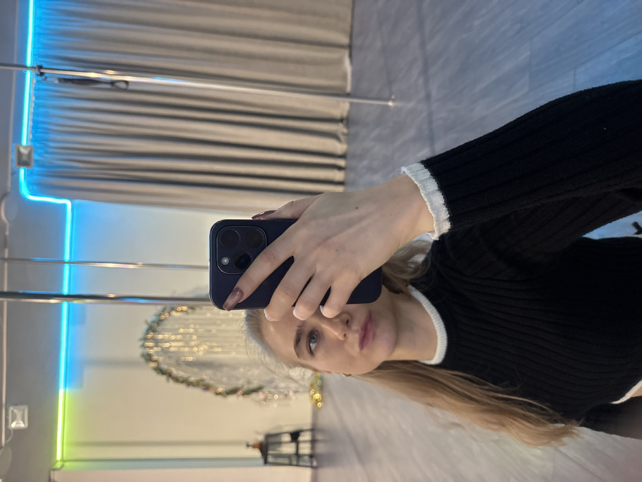 A woman takes a selfie in a mirror. She is wearing a black ribbed sweater with white trim around the neck and cuffs. She holds a dark blue phone with multiple camera lenses. The reflection shows a room with blue and green neon lights, curtains, and a decorative floral wreath. A small table and chairs are visible in the background.