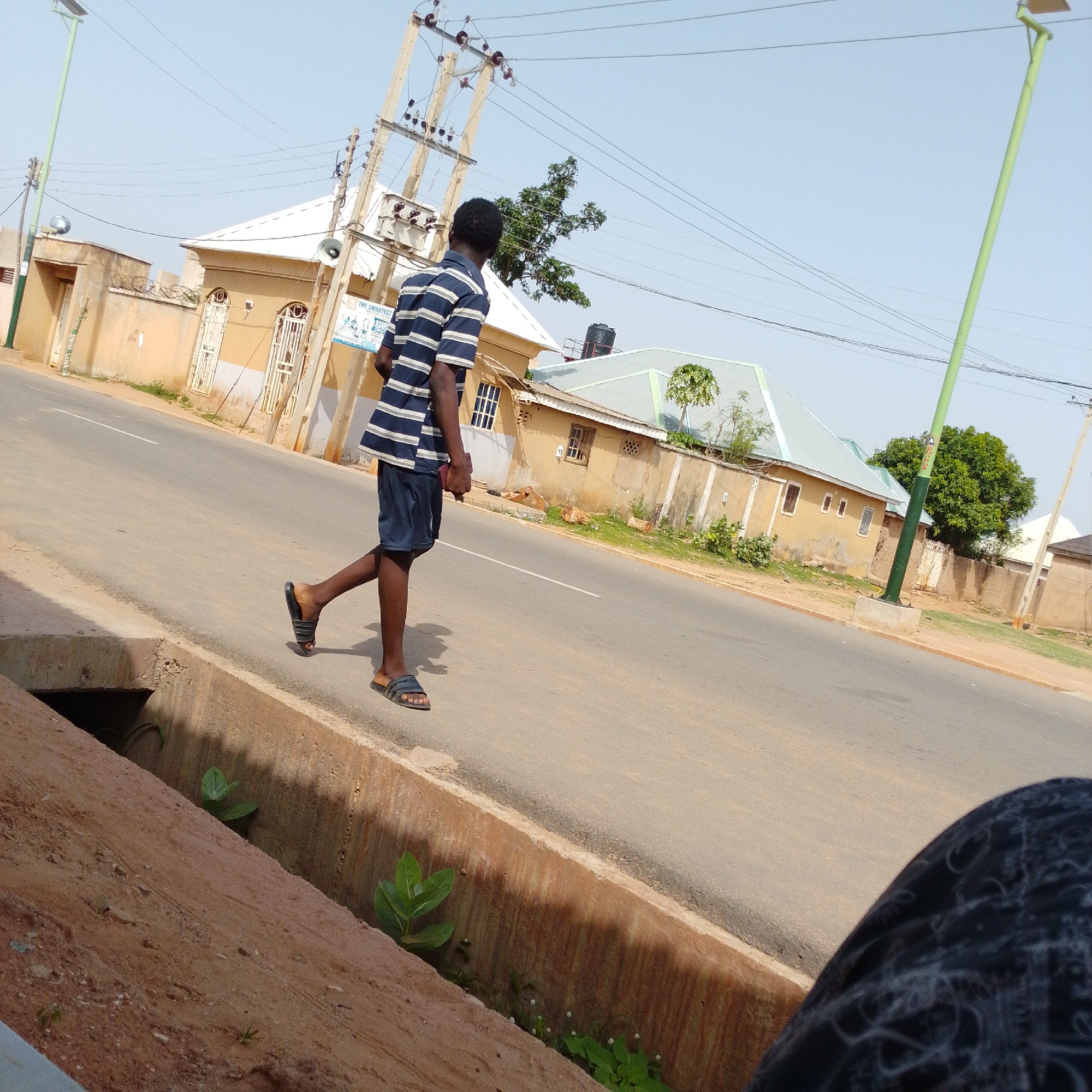 The image captures a street scene in Gombe, Nigeria, likely during daylight. A young man wearing a blue and white striped t-shirt and dark shorts walks on the right side of the paved road, his body angled away from the camera. He is wearing dark sandals on both feet. Beside the road, on the left, is a concrete drainage ditch with some green vegetation growing within it. Across the street, on the right, are several buildings with light-colored walls and metal roofs. Power lines and utility poles are visible overhead. The sky is clear and bright blue. The overall impression is of a typical urban environment in Gombe.