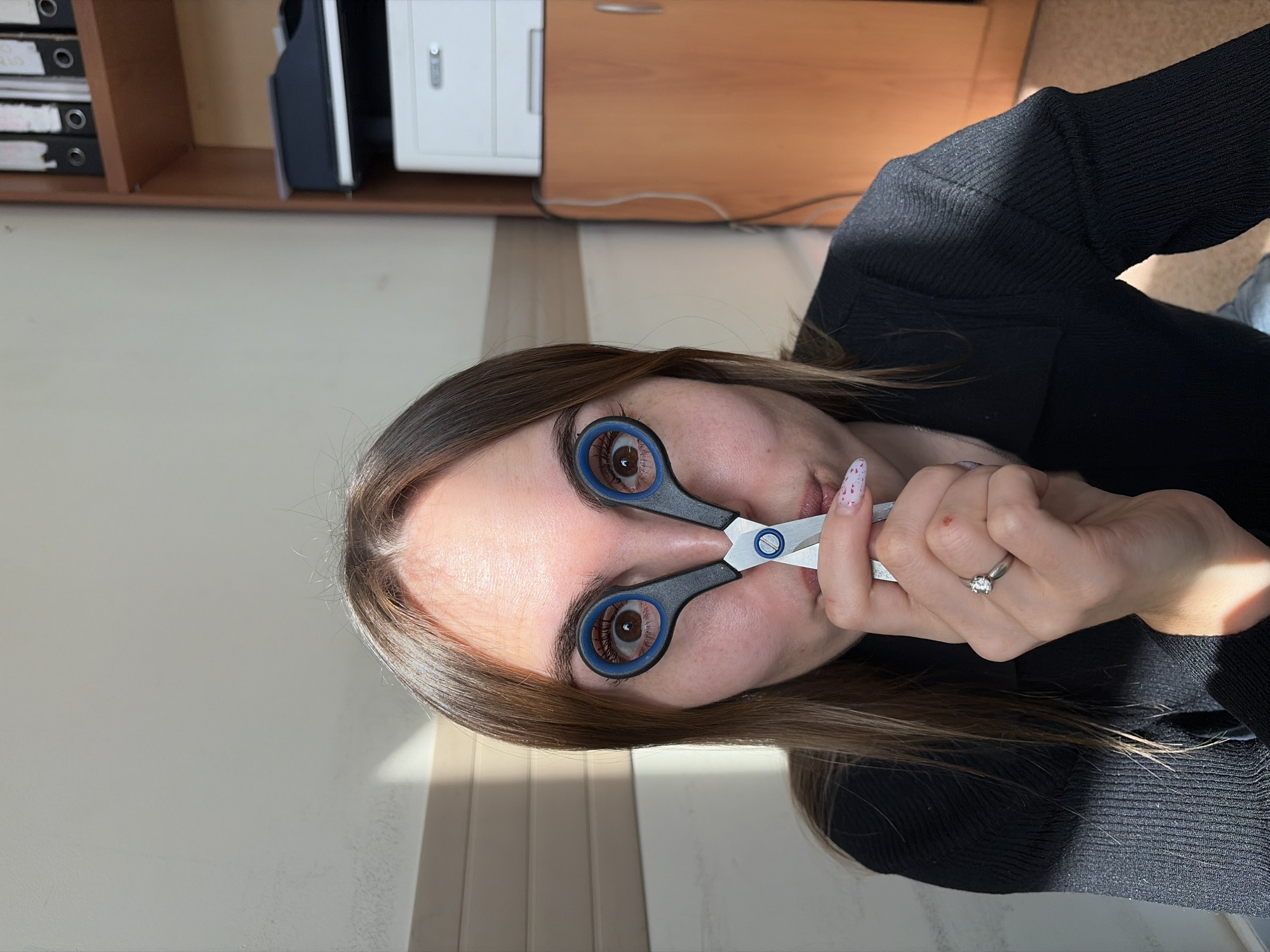 A fair-skinned female poses indoors, holding a pair of blue and white scissors in front of her face so that each eye is visible through a handle. Her hair is light brown and she is wearing a black sweater. She has a silver ring on the ring finger of her left hand. A white wall is visible to the left, with cabinets visible in the background. The location is Izhevsk, Russia.
