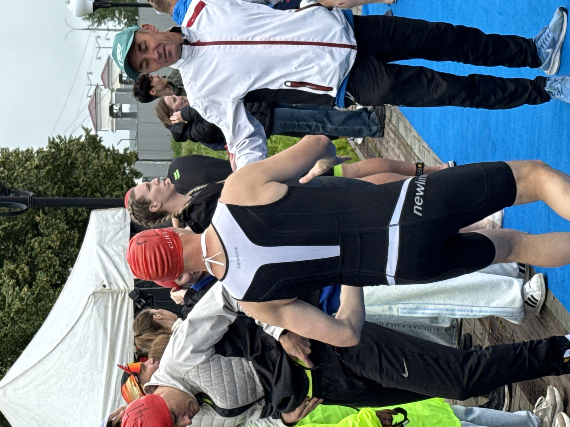 A group of athletes, some in racing gear, are gathered outdoors near a white tent, possibly at a sporting event in Votkinsk, Russia. A man in a white and red zip-up jacket and black pants, wearing a green cap, stands on a blue surface, facing to his right and appearing to speak with another man. The second man, facing away from the camera, wears a red swim cap and a black and white triathlon suit with the word "newline" visible on it. Several other individuals, dressed in athletic attire and casual clothing, are visible in the background, some under the tent and others standing around. The environment appears to be overcast, with trees and some distant buildings visible.
