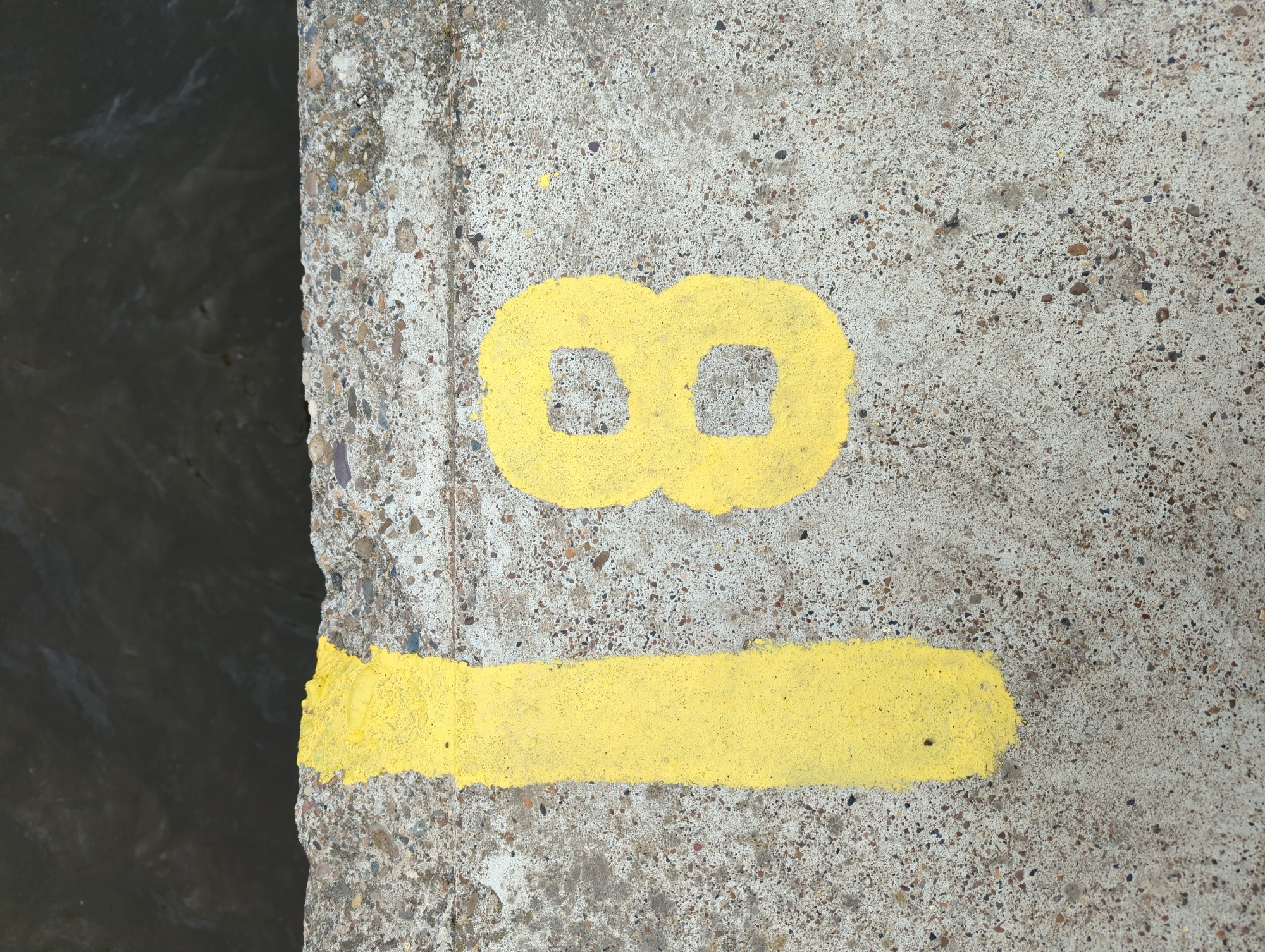 The image shows a close-up, overhead view of a concrete surface with the number "18" painted in yellow. The concrete is speckled with small stones. A dark, rippling body of water is visible in the upper portion of the image, separated from the concrete by a rough, uneven edge. The number "1" is on the left, painted as a thick, vertical stroke with a slightly irregular top. The number "8" is to the right, painted as two interconnected circles, also with some irregularity in the paint application. The scene appears to be outdoors, possibly at the edge of a dock or embankment. There are no people or animals visible in the image. The location of Kamskoye Ustye, Russia is not explicitly indicated by any visual cues in the image itself.