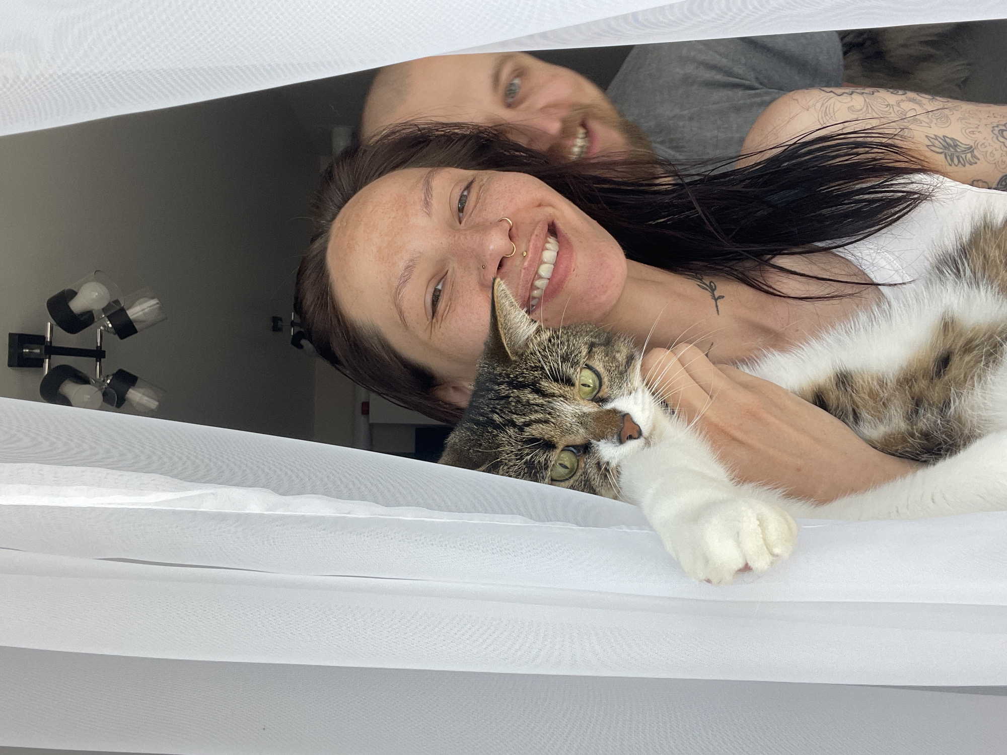 A medium shot depicts a woman, a man, and a cat posing behind a translucent white curtain. The woman is in the foreground, smiling, with dark brown hair. She is wearing a white tank top. The cat, a tabby and white mix, is in her arms, with its head near her face. Behind her, a man with a beard smiles, partially obscured. The background shows a dimly lit room. A light fixture is visible on the left. There is no information that places the image in Izhevsk, Russia.