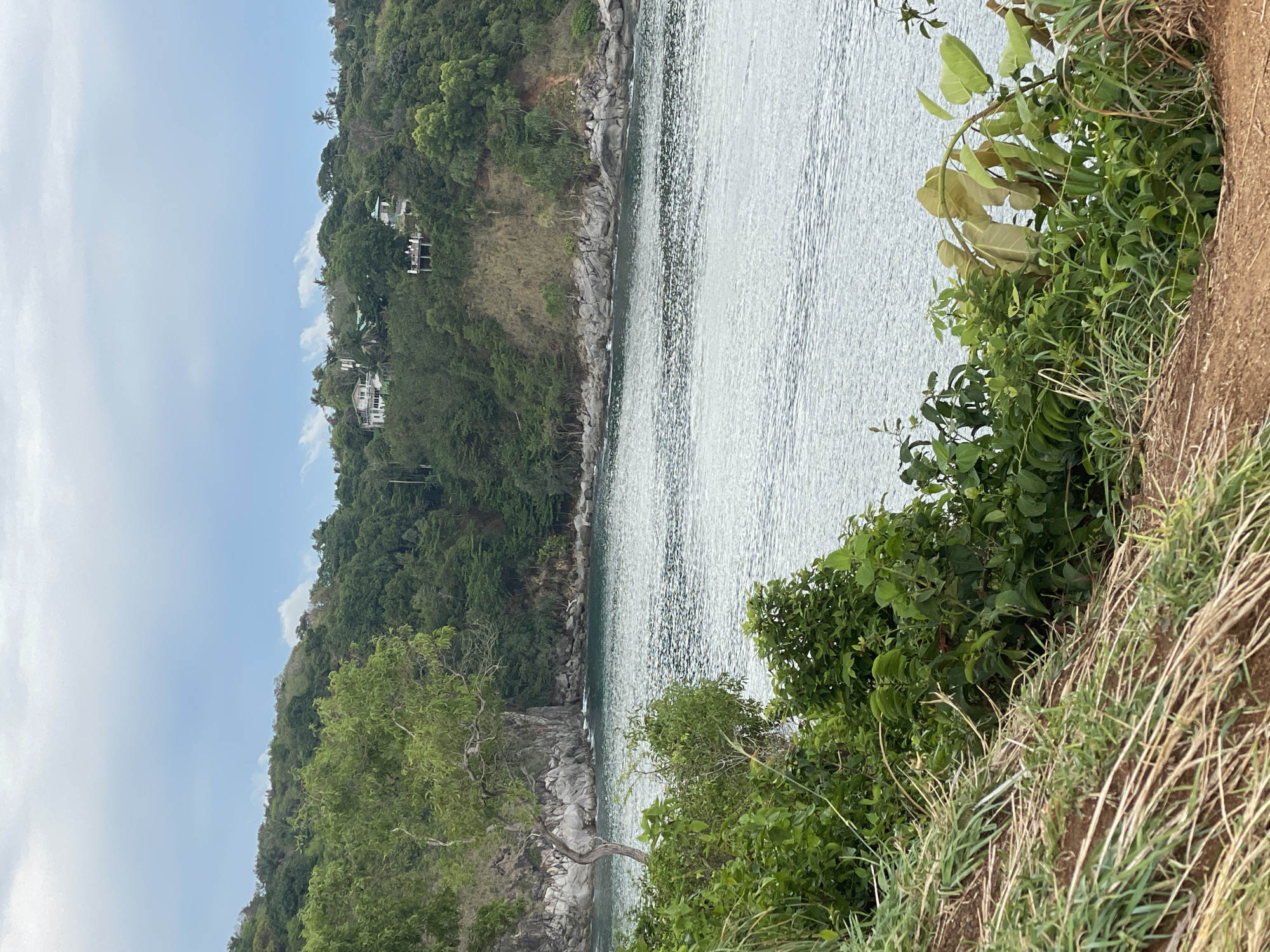 The image depicts a coastal scene in Rawai, Thailand. On the left, a steep hillside covered in dense tropical vegetation rises from the water's edge. Several buildings are visible among the trees. The sea occupies the majority of the frame. In the foreground, vegetation grows along the edge of a cliff or bank.