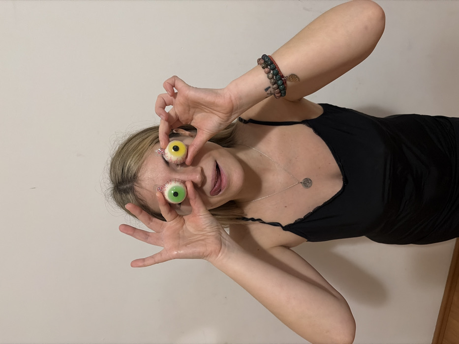 A woman in a black tank top playfully holds two wrapped candy eyeballs in front of her eyes. One eye is yellow, and the other is green. She has her tongue sticking out and her fingers splayed around the "eyes" for emphasis. She is wearing a silver necklace, and multiple bracelets on her arm. The background is a plain white wall, and the photo is rotated slightly.