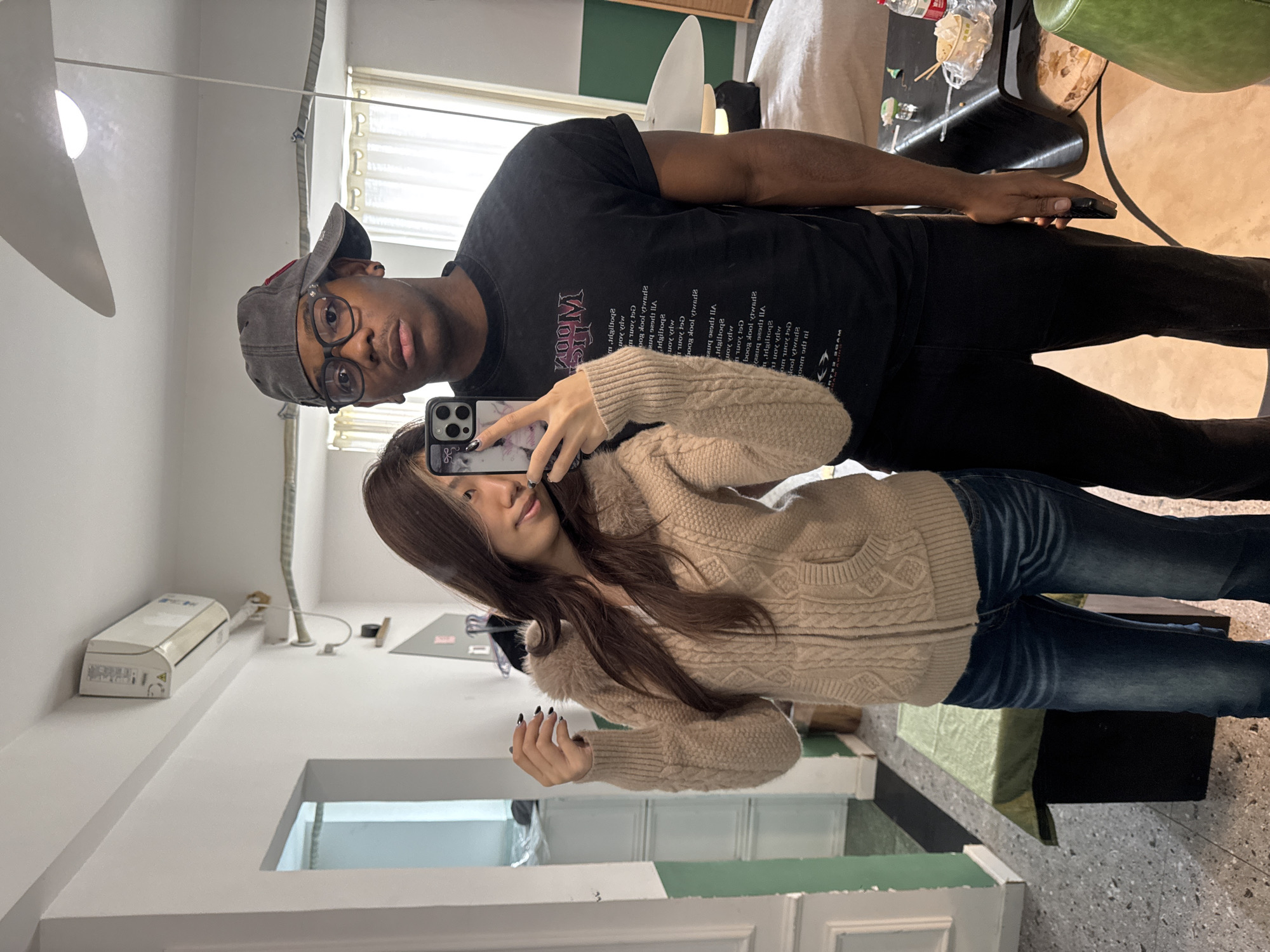 A mirror selfie captures two individuals within an indoor establishment in Liangjiang, China. In the foreground on the left, a female of East Asian ethnicity holds a smartphone with a black, floral/animal-patterned case, positioning it to take the photograph. She has long brown hair and is dressed in a beige cable-knit cardigan with a faux fur collar and blue jeans. Her left hand displays black-painted fingernails, and she has a slight smile, looking towards the camera. Standing behind and to the right of the female is a male of Black ethnicity. He wears a grey baseball cap backward, black-rimmed glasses, a black t-shirt with reversed white and pink text (reading "MOON" and a list of phrases), and black trousers. He looks directly at the camera with a neutral expression, his right hand holding a dark object down by his side.
The environment features white walls, with a portion of a doorway revealing a green-painted accent wall on the far left. The floor transitions from a speckled grey terrazzo-like material on the left to a lighter, possibly beige, surface on the right. A white air conditioning unit is mounted high on the wall in the upper left. White curtains cover a window in the upper right background, and a white ceiling fan is partially visible above the male's head. Furniture includes a green-upholstered bench or table on the far left, a light-colored padded bench or sofa on the right, and a black, curvilinear coffee table in the mid-right. On the coffee table, a red beverage bottle, a white plastic food container, chopsticks, and a small glass with a green element are visible. A green curved stool is partially visible in the bottom right corner.