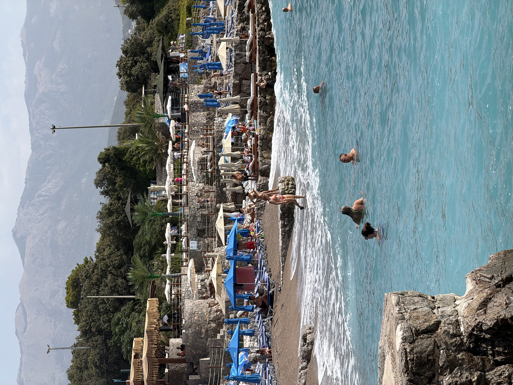 The image depicts a lively beach scene at a resort, likely located in Prijevor, Montenegro. The foreground features a rocky outcrop on the left, with vibrant turquoise water stretching out to the horizon. Several individuals are visible swimming in the water, including two women with blonde and dark hair respectively, and two men, one with his head above water and another further back. A person is wading into the water near the shore.
The middle ground showcases a sandy beach lined with rows of lounge chairs and numerous blue and white umbrellas, indicating a well-equipped recreational area. A few people are seen relaxing on the chairs or walking on the beach. A woman in a pink bikini walks at the edge of the water.
The background reveals a tiered resort complex built into a rocky hillside. Stone walls, thatched-roof structures, and more lounge chairs and umbrellas are visible. Dense green foliage and palm trees are interspersed throughout the resort. In the far background, majestic, hazy mountains rise against a pale sky, suggesting the mountainous terrain characteristic of the region. Streetlights are present in the resort area.