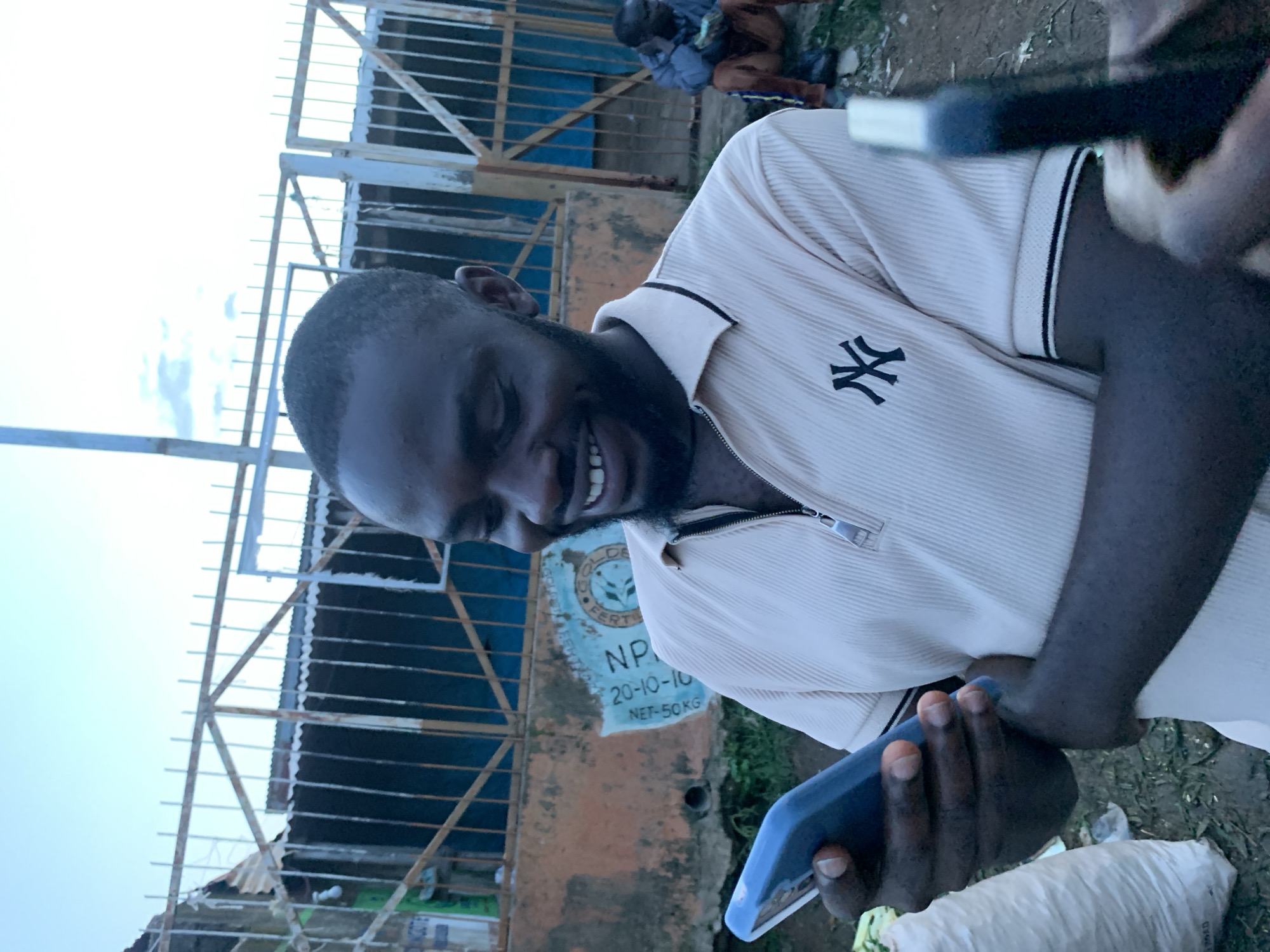 A young Black man is looking at his smartphone with a wide smile. He is wearing a light beige polo shirt with a black and white striped collar and a black NY logo on the left chest. His arms are crossed and he is holding the phone in his left hand. In the background, there is a rusty metal fence and a wall with a faded sign that appears to be for fertilizer. Behind the main subject, a second man is sitting on the ground, facing away from the camera. The environment appears to be outdoors, possibly in a market or street setting in Gura Top, Nigeria, indicated by the presence of bags of goods and the overall urban landscape. The sky is light blue with some clouds.