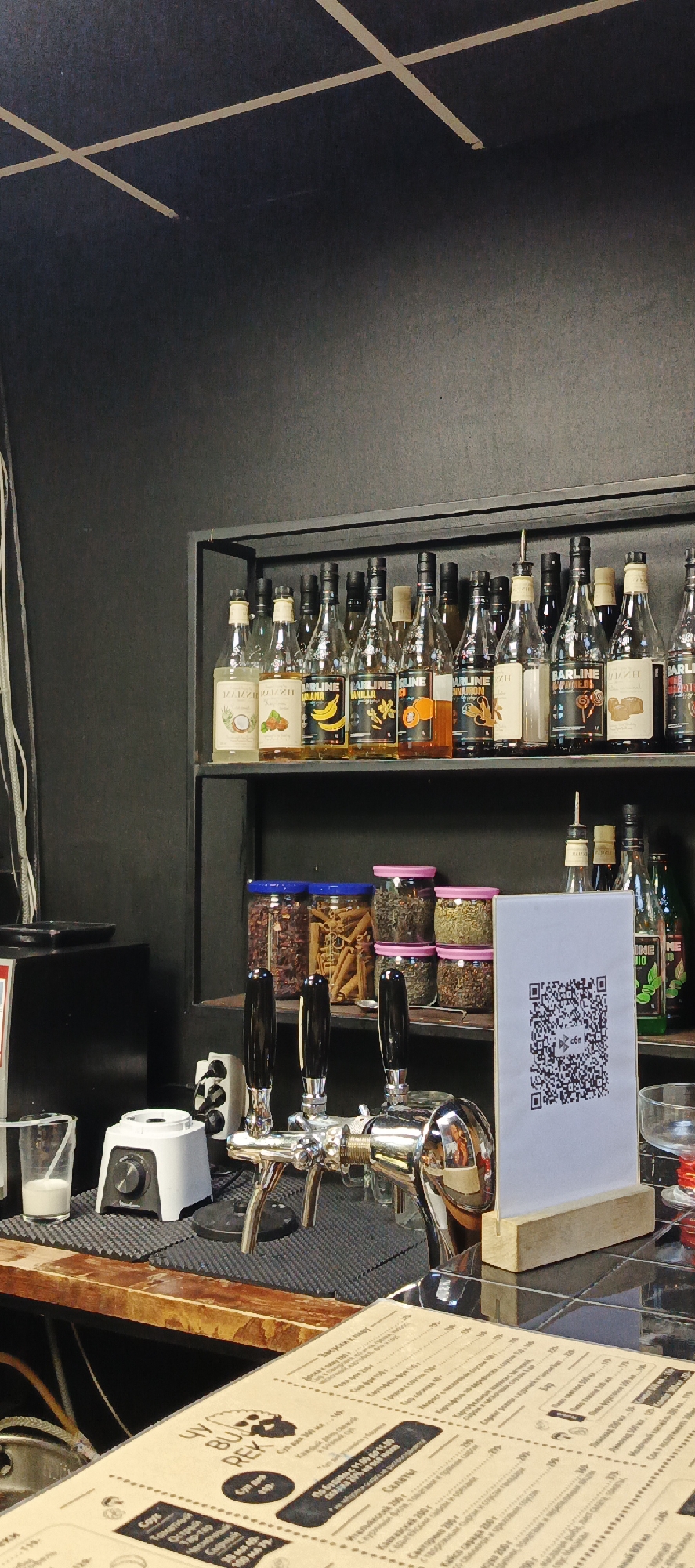 The image depicts a bar counter area with shelves of bottles and jars in the background. On the counter, there is a beer tap system with three handles, a small white appliance that appears to be a blender, and a clear plastic cup with a white liquid. To the right of the tap system is a white stand with a QR code on it. The shelves in the background are filled with various bottles of syrups or liqueurs, some with labels indicating flavors like coconut, hazelnut, banana, vanilla, and caramel. There are also several glass jars containing what appear to be dried goods like spices or tea. The bar area is dimly lit with a dark, textured wall in the background and a wooden countertop. There are no people visible in the image. The overall scene suggests a bar or cafe setting. The presence of the QR code indicates a modern business that likely offers digital payment or menu options. Based on the context of Votkinsk, Russia, this scene is likely from a local establishment.