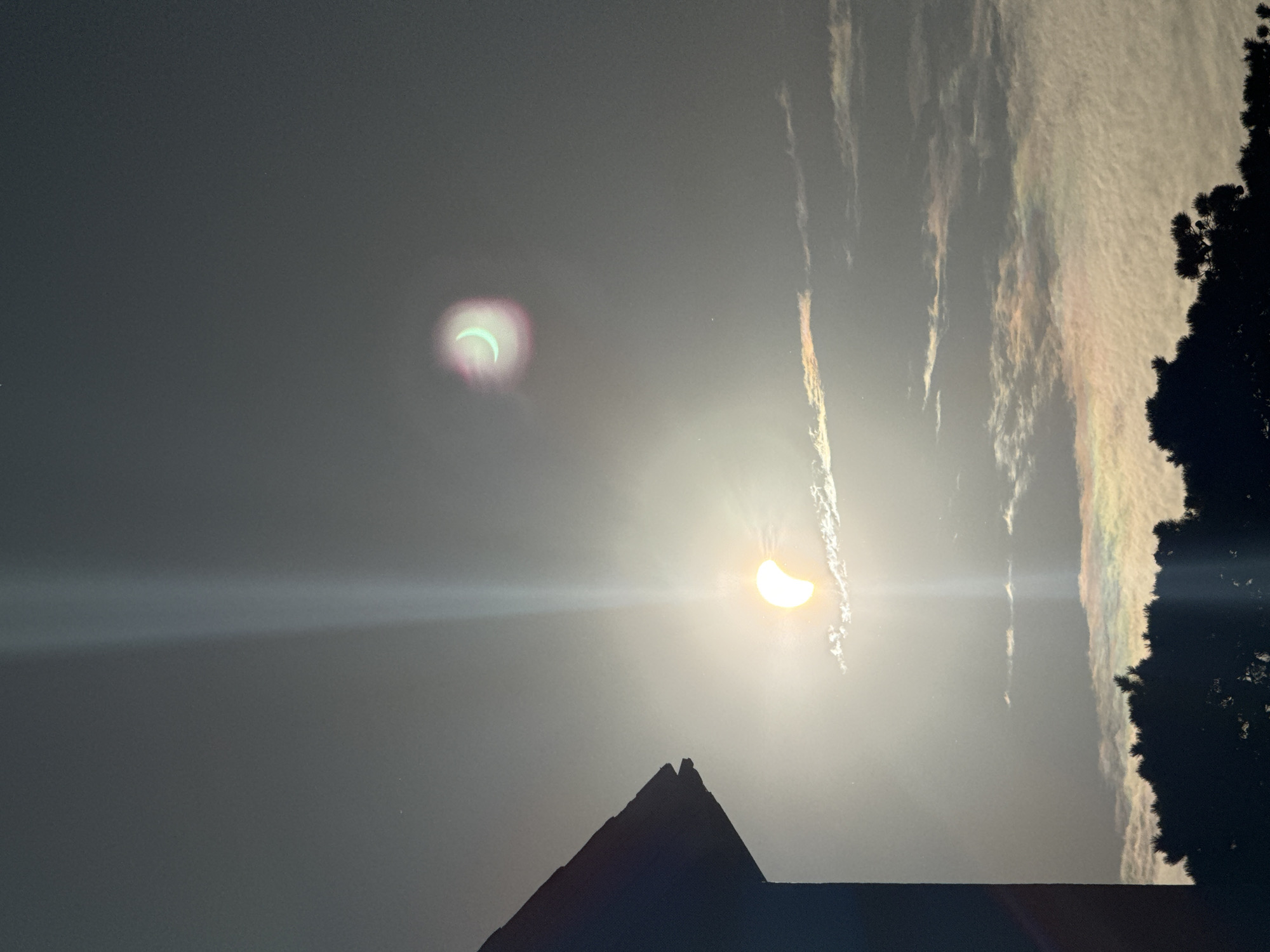 The image depicts a partially eclipsed sun viewed from Waterford, Ireland. The sun, visible as a crescent shape, is partially obscured by the moon. There is a prominent light flare extending vertically through the image. In the upper right portion of the sky, a second, smaller crescent-shaped object, possibly another solar phenomenon or artifact, is visible with a greenish hue surrounded by a pinkish halo. The foreground shows the silhouette of a building on the left and the dark outline of trees at the bottom. Wispy clouds are present in the sky, some reflecting iridescent colors, indicating a potential atmospheric phenomenon like iridescence. The overall scene is bathed in the bright light of the sun, creating a hazy and atmospheric effect.
