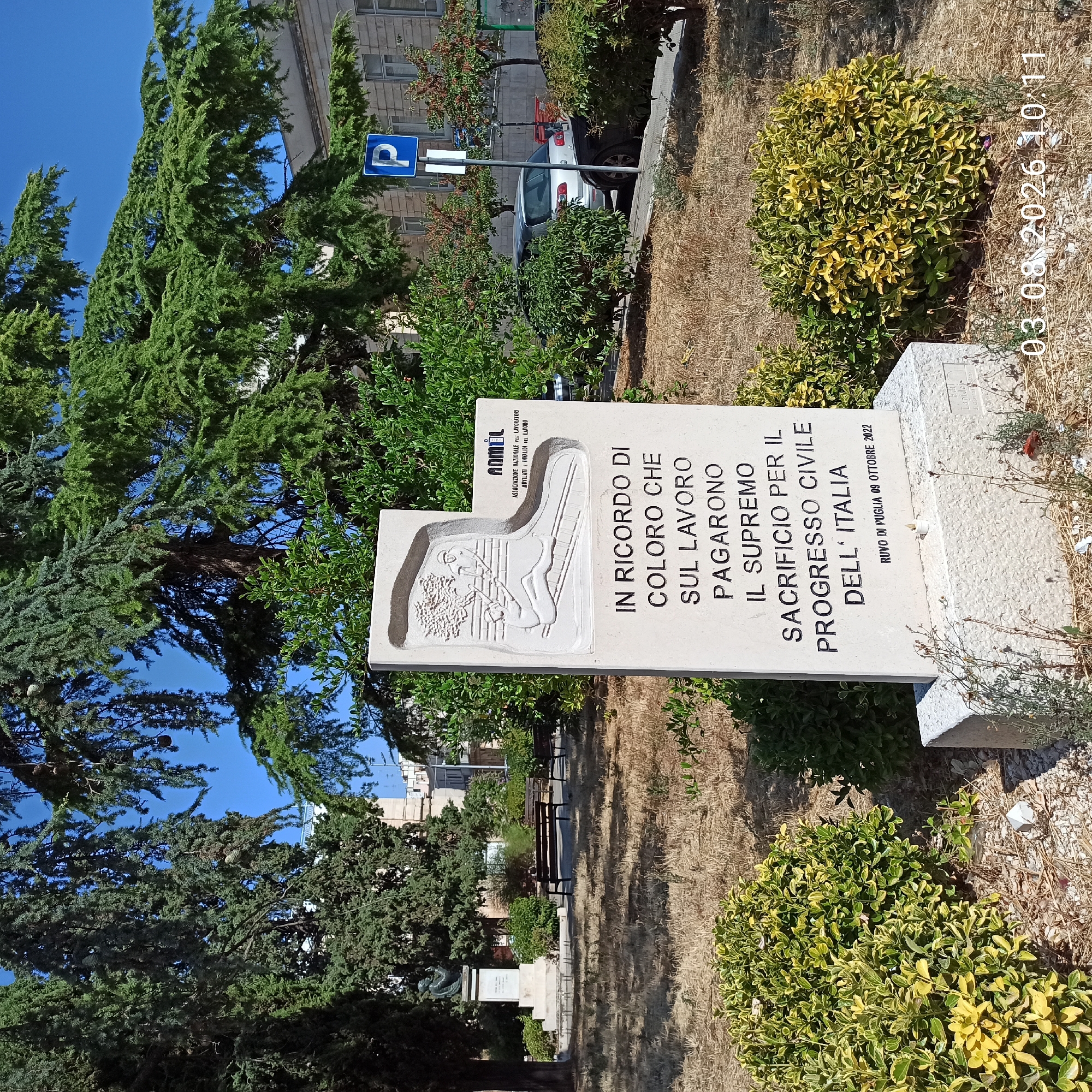 The image depicts a memorial monument in an outdoor setting, likely a park or public square, in Ruvo di Puglia, Italy. The monument, made of light-colored stone, features an inscription in Italian and a bas-relief carving. The inscription reads, "IN RICORDO DI COLORO CHE SUL LAVORO PAGARONO IL SUPREMO SACRIFICIO PER IL PROGRESSO CIVILE DELL' ITALIA," which translates to "In memory of those who on work paid the supreme sacrifice for the civil progress of Italy." Below this, it states, "RUVO DI PUGLIA 09 OTTOBRE 2022." The bas-relief shows two figures engaged in labor, possibly construction or manual work, with a stylized tree in the background.
The monument stands on a textured concrete base. To the left of the monument, a portion of a classical-style sculpture on a pedestal is visible, suggesting a historical or civic significance to the location. The surrounding environment includes dry, yellowed grass, several green bushes, and large evergreen trees with a clear blue sky above. In the background, a building with a light-colored facade and windows can be seen, along with a blue parking sign with a white "P". A white and silver car is parked on the side of a paved area to the right of the monument, and other foliage and trees are scattered throughout the scene. The overall atmosphere is one of solemn remembrance and civic pride.