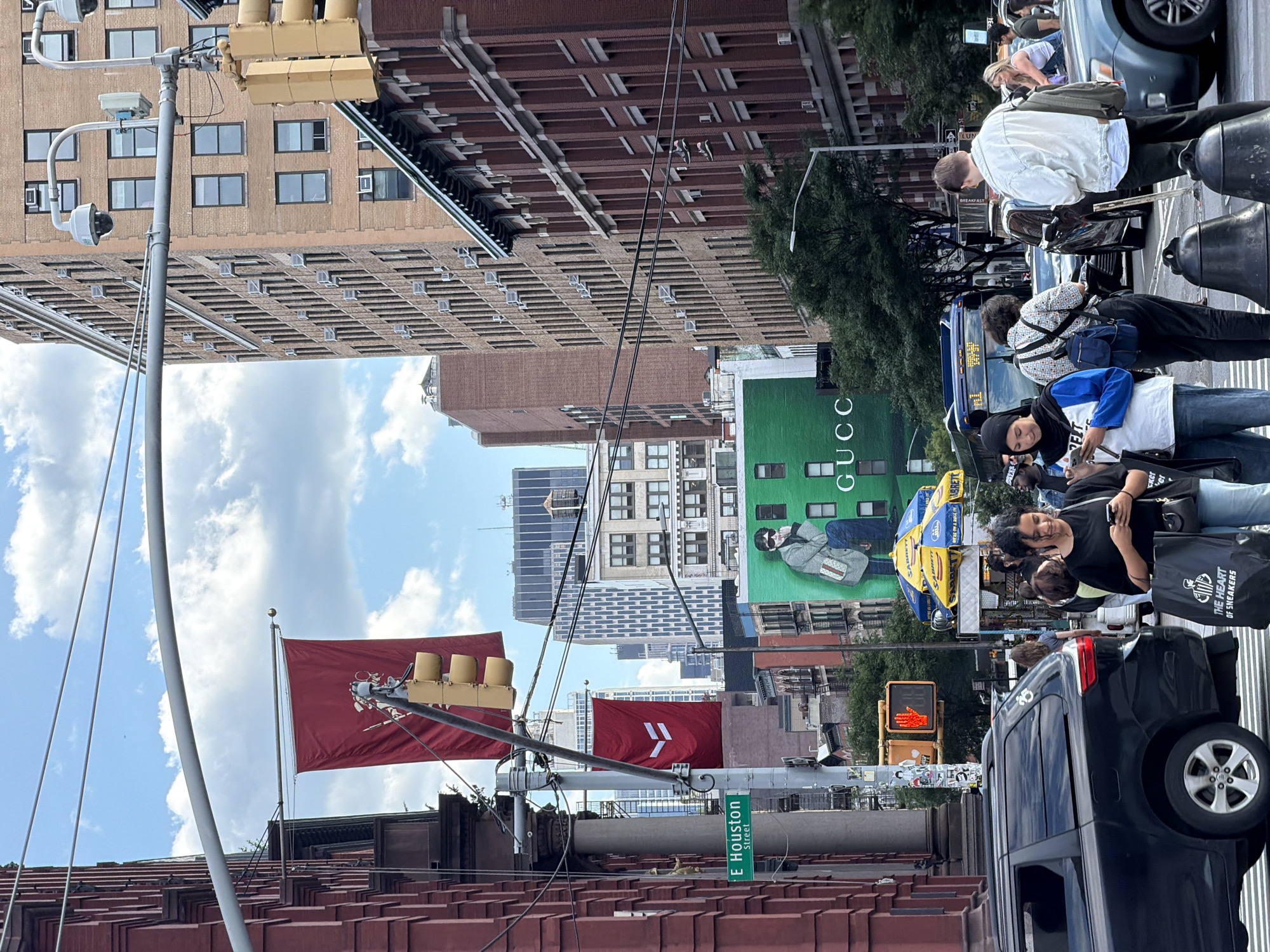 The image depicts a street scene in New York City. Pedestrians are crossing the street, and vehicles are visible. There are tall buildings in the background, including one with a large "GUCCI" advertisement. Street signs indicating "E Houston Street" are present. A food vendor is visible as well as pedestrians with shopping bags. Flags on buildings are also visible. The sky is partly cloudy.