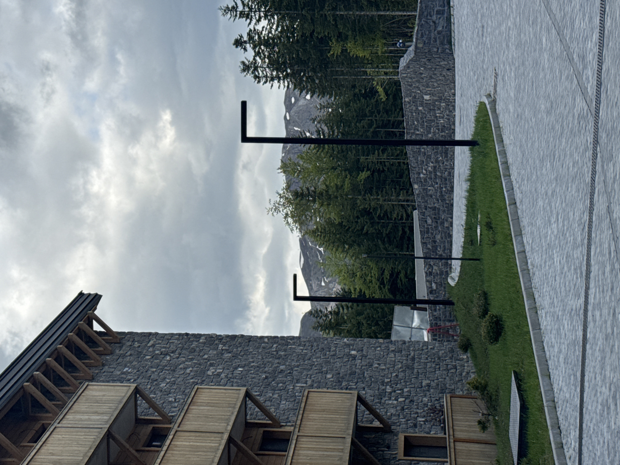 The image shows a mountainside landscape in Katun Bačko brdo, Montenegro, on an overcast day. A modern stone building with wooden accents is in the foreground. Tall, thin evergreen trees, along with a rocky mountain peak with snow, are behind the building. Several black, L-shaped lampposts are between the building and trees. A paved path with a strip of green grass is alongside a stone wall. The sky is covered in gray clouds.