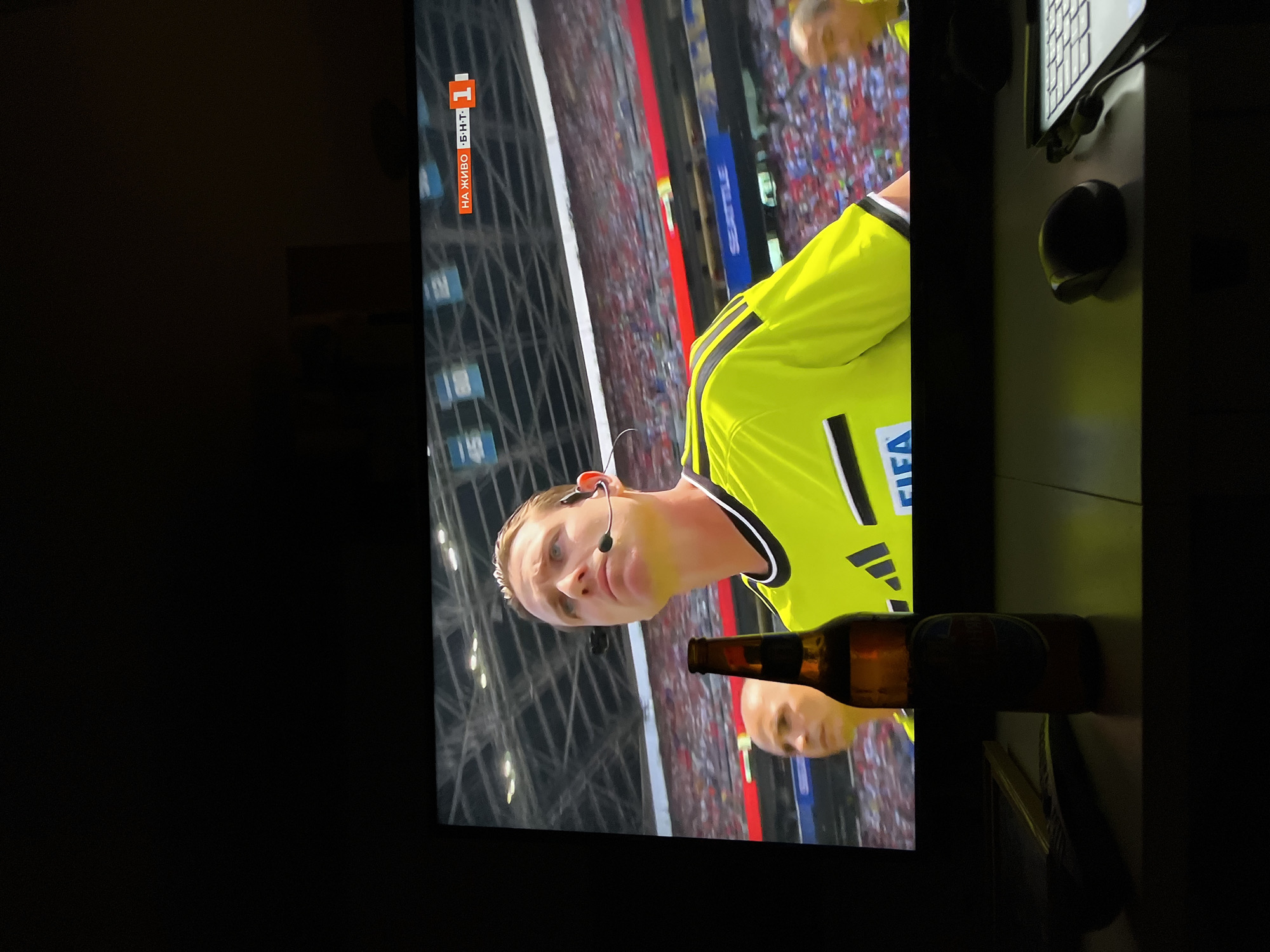 The image displays a television screen in a dimly lit room, showing a soccer match. A referee in a bright yellow jersey with black trim and a microphone headset is visible in the foreground on the screen. To his left, another man in a similar uniform is partially visible. The background of the screen depicts a stadium filled with spectators. In the top right corner of the screen, a graphic indicates "НА ЖИВО Б.Н.Т. 1", suggesting the broadcast is live on BNT 1.
In front of the television, on a desk, a partially consumed bottle of beer stands upright. To the left of the beer bottle, a dark object, possibly a remote control or a phone, is placed. Further to the right on the desk, a computer mouse and part of a keyboard are visible. The setting appears to be a home environment, likely in Sofia, Bulgaria, given the presence of the Bulgarian national television channel graphic. The overall scene captures a moment of watching a sporting event with a drink and typical desk accessories.