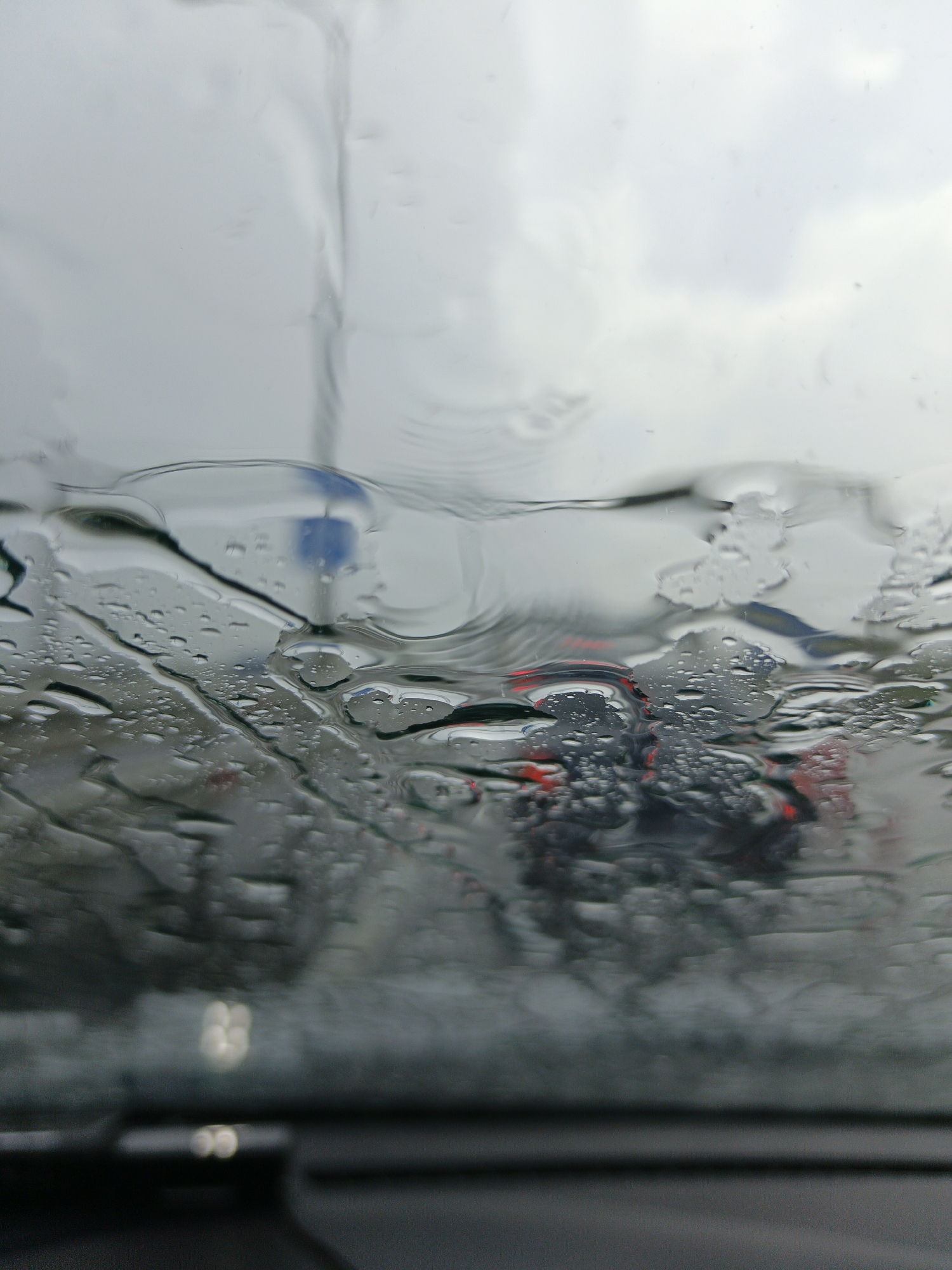 The image depicts a view through a rain-streaked car windshield, looking out at a street scene in Kerch, Ukraine. The dominant elements are the water droplets and streaks on the glass, distorting the view of the environment beyond. In the mid-ground, what appear to be the taillights of vehicles are visible as red blurs, suggesting traffic. A blue traffic sign is also discernible through the water, though its details are obscured. The sky is overcast, contributing to the muted, gray tones of the scene. The foreground shows a portion of the car's interior, including a dark dashboard and some indistinct reflections. The overall impression is one of a rainy day commute or stop-and-go traffic in an urban setting.