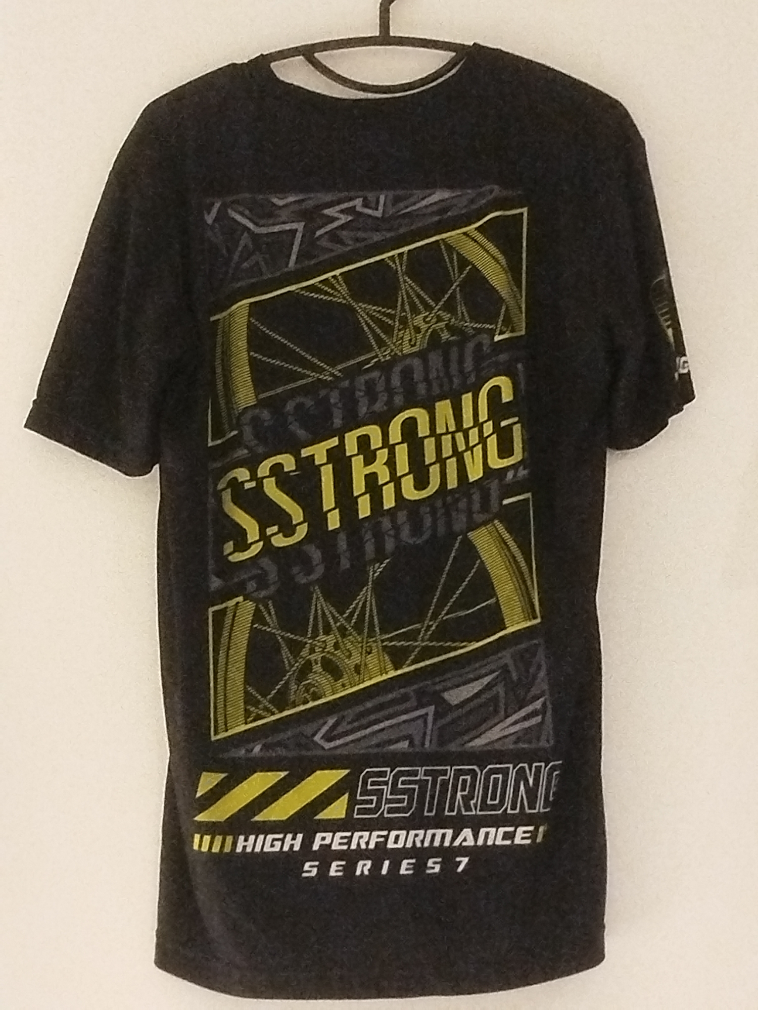 The image shows a dark gray t-shirt hanging on a white wall. The t-shirt has a graphic design on the front featuring yellow and gray elements. The design includes stylized bicycle wheels with spokes, and repeated text that appears to say "SSTRONG". Near the bottom of the graphic, there is text that reads "HIGH PERFORMANCE" and "SERIES 7". The t-shirt is on a black hanger. The lighting suggests an indoor setting. No people are visible in the image. The location is not discernible from the image itself.