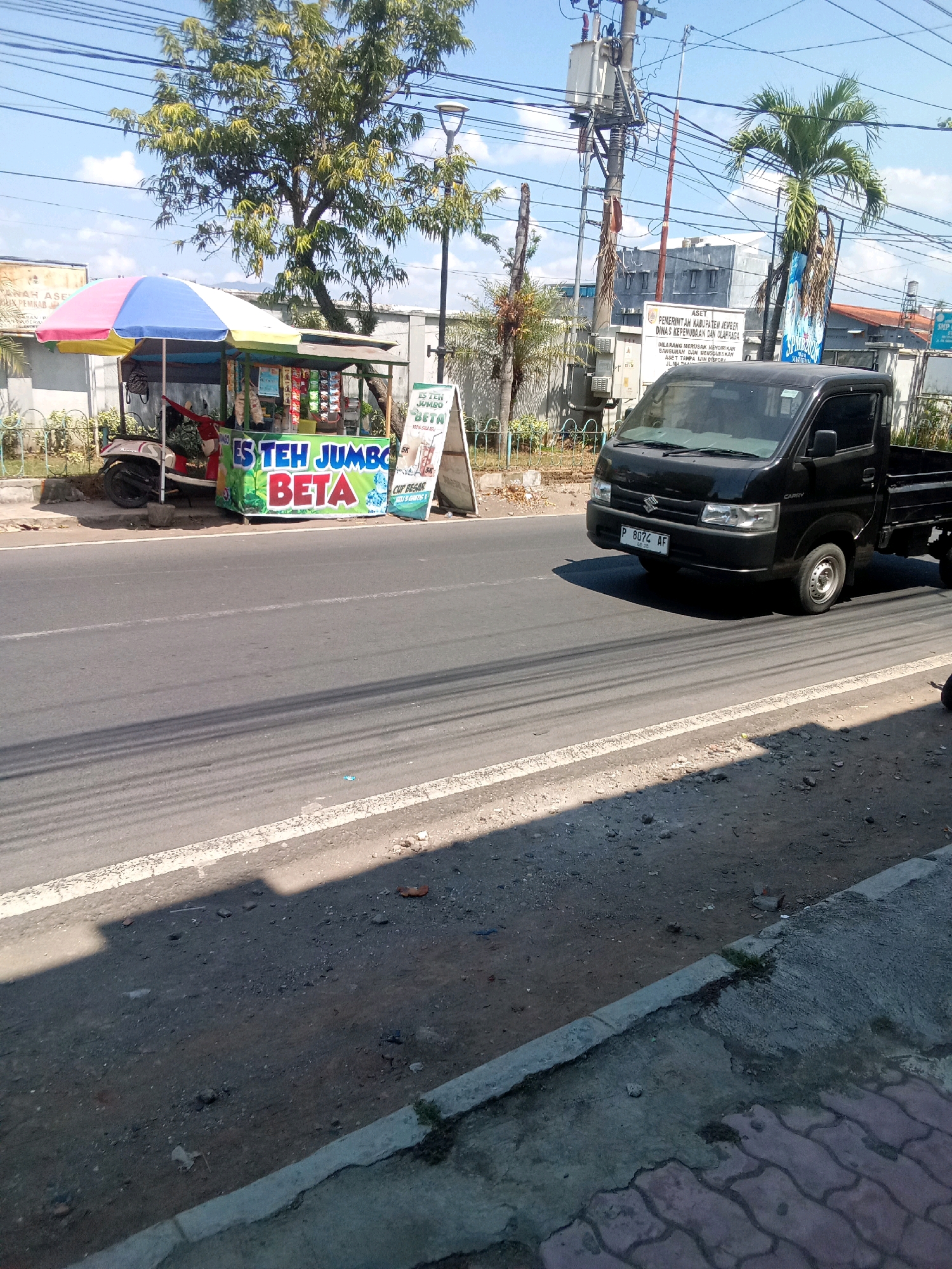 This image depicts a street scene in Jember Kidul, Indonesia, likely on a sunny day. A black Suzuki Carry pickup truck is driving on the right side of the road, with its license plate visible as P 8074 AF. On the left side of the road, a small food cart is set up under a colorful umbrella. The cart appears to be selling drinks, with signage that reads "ES TEH JUMBO BETA". A red scooter is parked behind the cart. In the background, there is a building with a sign that reads "PEMERINTAH KABUPATEN JEMBER DINAS KEPENDUDUKAN DAN CATATAN SIPIL" which translates to "JEMBER REGENCY GOVERNMENT DEPARTMENT OF POPULATION AND CIVIL REGISTRATION". There are also power lines and trees visible. The road surface is asphalt with white lane markings. The surrounding area appears to be an urban or semi-urban environment.