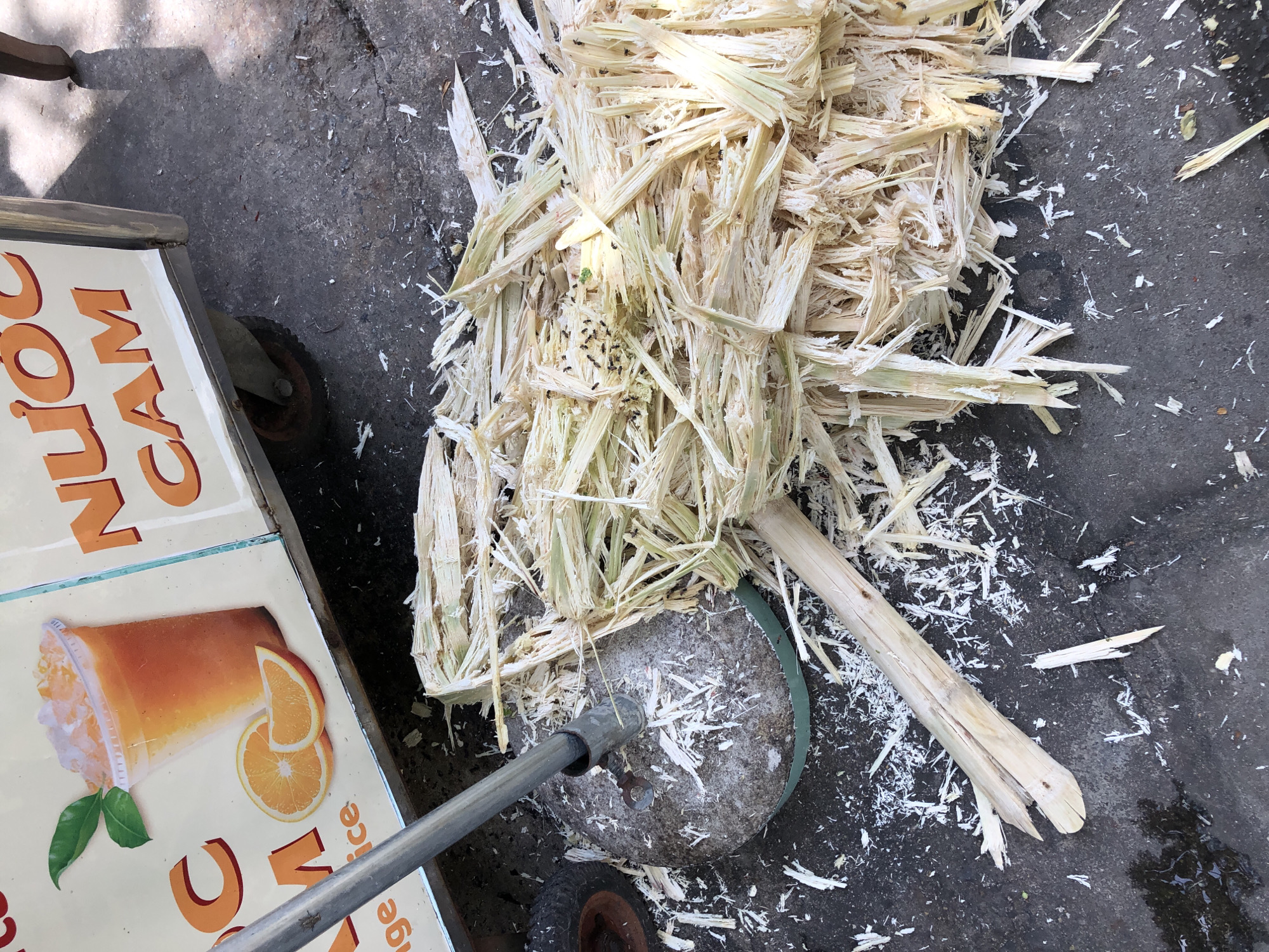 An outdoor scene in Da Nang, Vietnam, shows a pile of shredded sugar cane fibers on a dark asphalt surface. The sugar cane appears to have been processed by a nearby machine, indicated by a metal pole extending from a round, concrete base with a green rim, located to the left of the pile of fibers. Scattered white fragments of sugar cane are visible on the ground around the machine and the pile. In the background, part of a food cart is visible, with signage featuring text in Vietnamese and an illustration of a glass of orange juice with orange slices and leaves. The cart has orange lettering that reads "NƯỚC CAM." The overall impression is one of a street vendor's workspace where fresh juice is prepared.