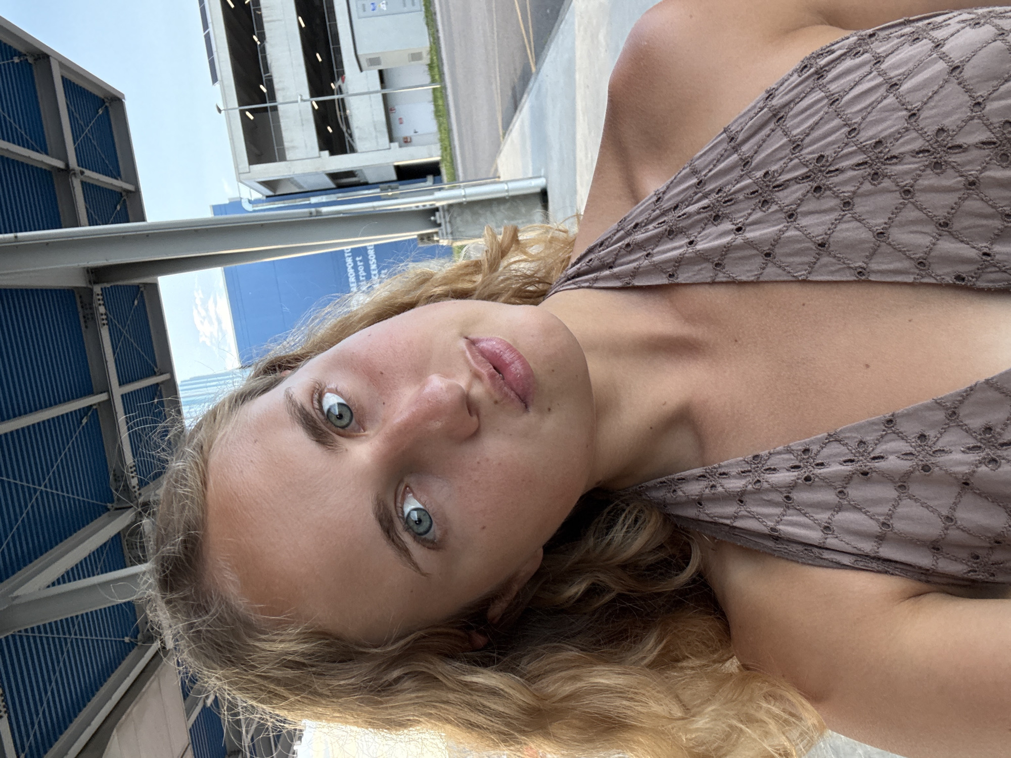 A young Caucasian woman with blonde, curly hair is taking a selfie outdoors. She is looking at the camera with blue eyes and her head is tilted to the side. She is wearing a light brown halter top with a diamond pattern. In the background, there is a blue building with text that appears to say "AEROPORTO" which translates to "Airport", and a multi-story parking garage. The setting is likely the Trieste Airport, located in Ronchi dei Legionari, Italy.