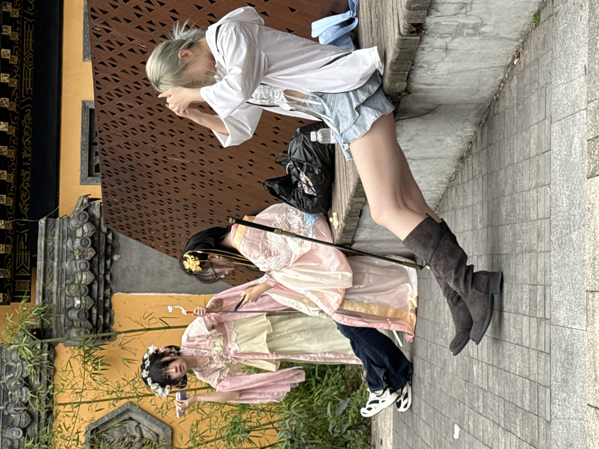 A daytime scene in Yuzhong District, China, captures three individuals amidst a blend of traditional architecture and urban elements. The image is oriented vertically.
In the upper portion, a person with light-colored hair, dressed in a white long-sleeved shirt, light blue denim shorts, and dark knee-high boots, leans forward, holding a phone or camera to take a picture. A light blue face mask is visible hanging from their attire, and a black plastic bag is at their feet.
Below, two individuals are dressed in elaborate light pink traditional Chinese garments, possibly Hanfu, with modern accents. The person in the middle has dark hair adorned with golden hairpins and holds a long, thin object resembling a sword or staff, pointing downwards. The person positioned further down also has dark hair, wearing a flower crown, and holds a small, rectangular device, possibly a phone or camera, while looking towards the middle person. Both individuals in traditional dress are notably wearing dark blue jeans and white and black athletic shoes beneath their robes, suggesting a modern interpretation or blend for a photoshoot.
The background on the left features traditional Chinese architectural elements, including ornate black and gold patterns, gray tiled roofs, and yellow walls, partially obscured by bamboo plants. On the right, a reddish-brown wall with a distinctive geometric cutout pattern serves as a backdrop. The ground is a paved stone walkway with visible cracks and some scattered debris. The overall activity appears to be a cultural photography session, combining historical aesthetics with contemporary fashion. No visible text is present in the image.