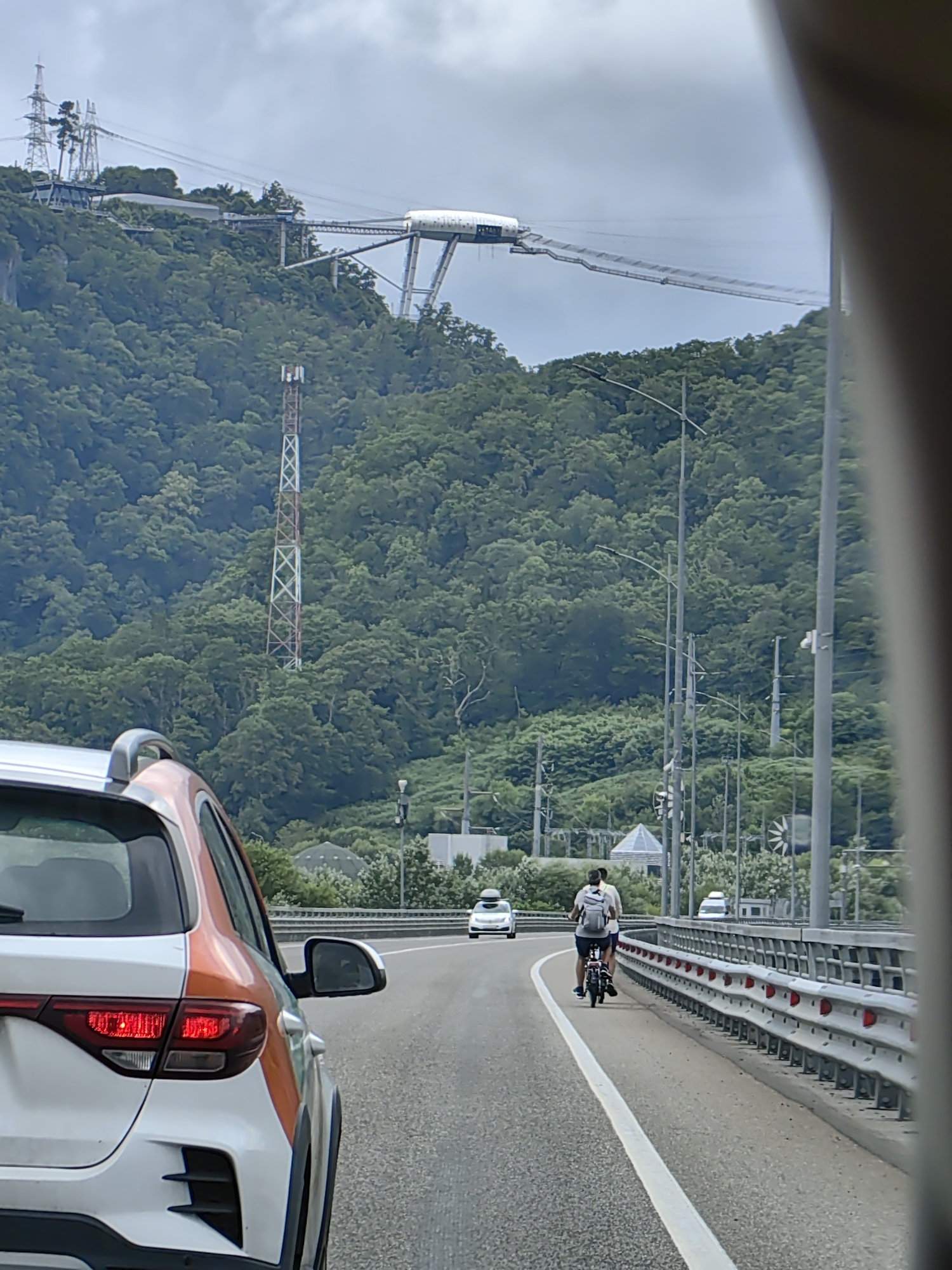 This is a photograph taken from a vehicle on a bridge, looking towards a lush, green mountainside under a cloudy sky. In the foreground, the rear of a white and orange car is visible. Further down the road, a person on a small bicycle is riding towards the camera, wearing a backpack. Several other vehicles are visible on the road, including a white car further ahead and another white vehicle in the distance. The bridge has a metal guardrail with red reflectors. On the mountainside, there is a tall telecommunications tower and a distinctive aerial walkway or bridge structure spanning across the mountain peaks. The location is identified as Kepsha, Russia.