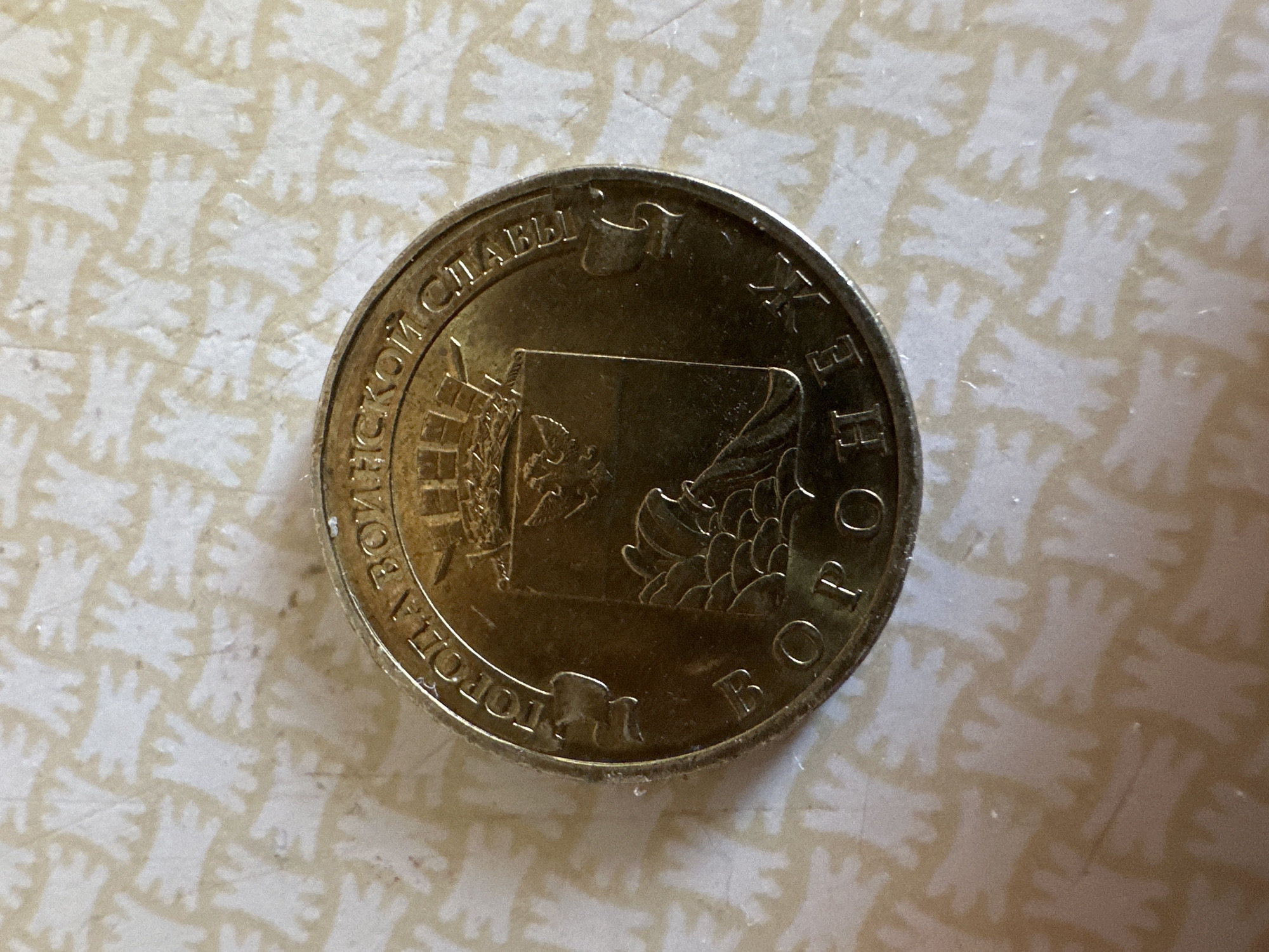 The image displays a close-up, overhead view of a single, circular coin. The coin is golden in color and appears to be made of metal. It is placed on a textured surface with a repeating pattern of light-colored, abstract shapes resembling human figures or stylized hands against a darker background.
The coin's surface features embossed text and an image. The text encircles the outer edge of the coin. The visible portion of the text includes Cyrillic characters. The inner part of the coin displays a coat of arms, which depicts a crown at the top, a shield with an eagle holding a sword and orb, and a landscape at the bottom featuring what appears to be hills or mountains and a stylized representation of a river or water.
The inscription on the coin reads "ГОРОДА ВОИНСКОЙ СЛАВЫ ВОРОНЕЖ" which translates to "City of Military Glory Voronezh". The city of Voronezh is a city in Russia, not Biysk. There is no information in the image to suggest any connection to the city of Biysk.