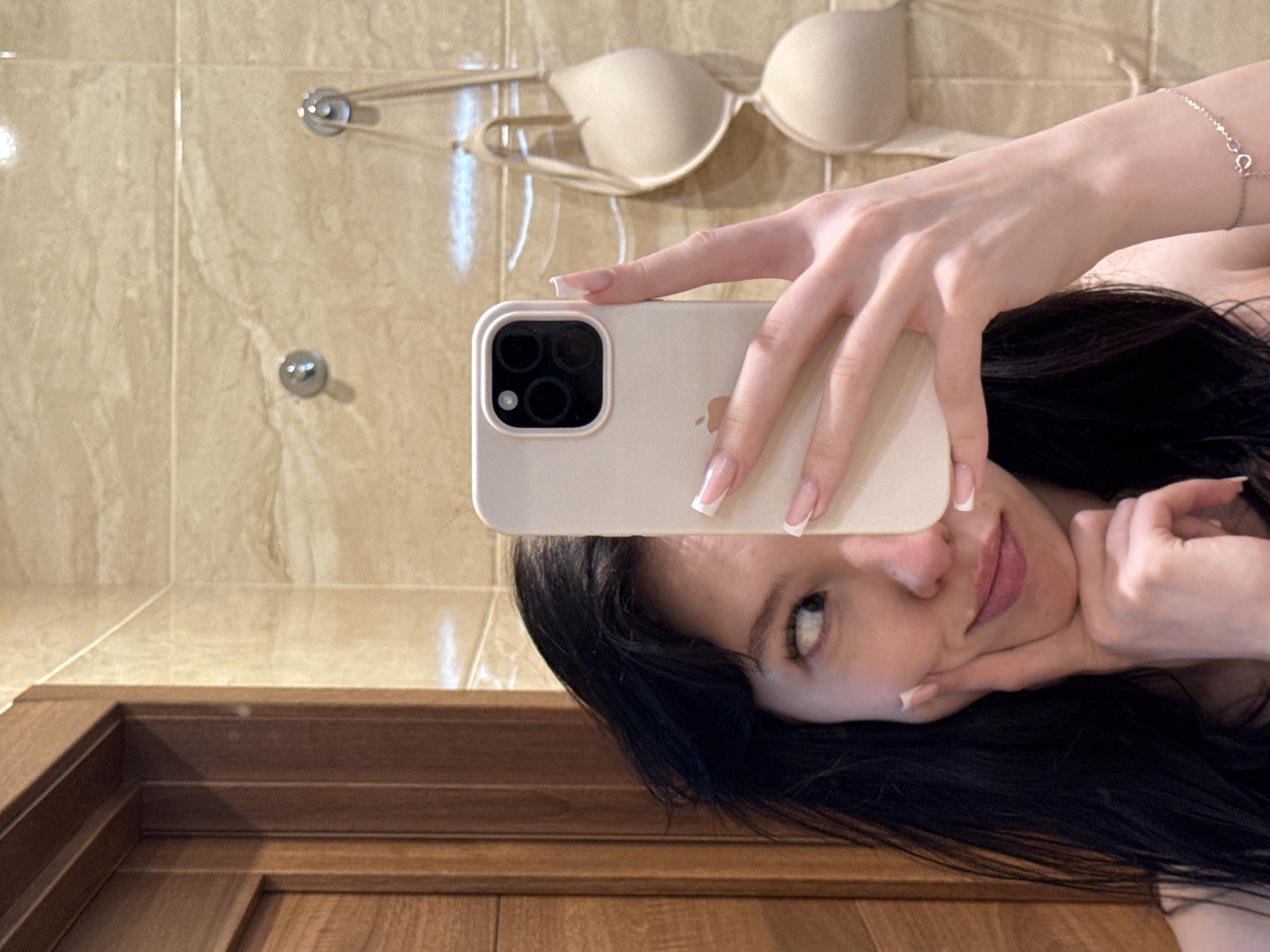 An indoor, eye-level, medium shot shows a young woman taking a selfie in a bathroom. Her face, partially obscured by a phone, is turned to her right, with her left eye looking off to the side. Her left hand rests on her chin, and her right hand holds up a white smartphone. She has dark, wavy hair and is wearing a thin-strapped top. In the background, a wall of tan marble tiles is visible, with a wooden door on the left. A beige bra is hanging on a hook on the tiled wall. The scene appears to be in a private residence in Khimki, Russia.