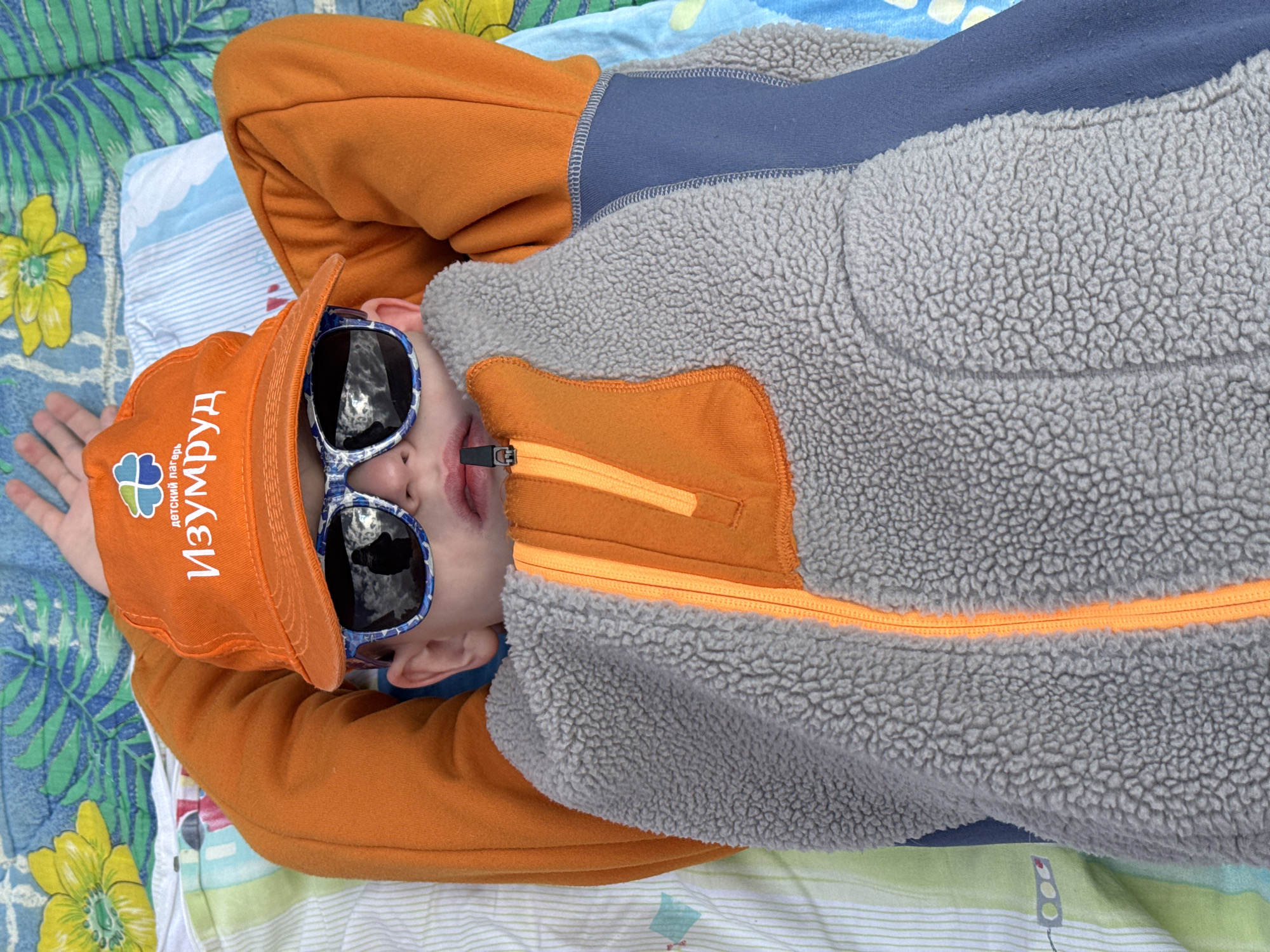 A child is lying on a bed or blanket outdoors. The location is unspecified. The child is wearing an orange baseball cap with Cyrillic text "Изумруд" which translates to "Emerald", and the text "детский лагерь" which translates to "children's camp". The cap also features a logo of a clover with a heart. The child is also wearing sunglasses and a gray and orange fleece jacket. The background is a patterned fabric with floral designs.