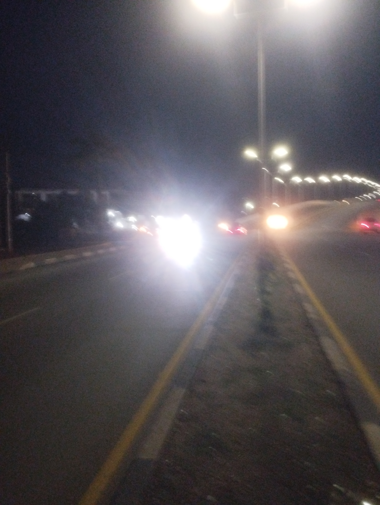 This is a night-time photograph taken in Jos, Nigeria. The image depicts a road with streetlights illuminating the scene. The primary subjects are vehicles, identifiable by their headlights and taillights, which appear as bright blurs due to motion and low light conditions. The road has yellow lane markings. In the background, there are some blurred structures that could be buildings or trees.
The activity taking place is traffic on a road at night. The bright lights suggest that it is dark, and the presence of streetlights indicates an urban or semi-urban environment. The weather appears clear, as there are no visible signs of rain or fog. The overall mood is somewhat obscured by the blurriness, but the illuminated road and moving vehicles suggest a normal evening flow of traffic. There is no visible text in the image.