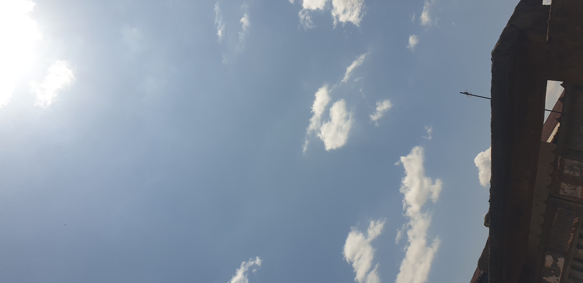 The image captures an outdoor scene in Sabon Tasha, Nigeria, dominated by a bright, partly cloudy sky. The sky, occupying approximately 75-80% of the frame, is a pale blue. The sun, manifesting as an intense white glare, is visible in the upper right quadrant, partially obscured by a thin, luminous cloud. Scattered white cumulus clouds are distributed across the mid-section and lower-left areas of the sky. A small, dark, unidentifiable object, possibly a bird or insect, is visible against the blue sky in the upper-left quadrant.
The lower 20-25% of the image features a dark, textured, horizontal structure, consistent with a weathered concrete wall or parapet, extending across the foreground. This structure exhibits a dark brown-grey color and appears in silhouette. Behind this foreground element, sections of reddish-brown corrugated metal rooftops are discernible, indicating the presence of other buildings. On the right side of the foreground structure, a slender, dark, vertical pole or antenna, possibly with minor horizontal components, extends upwards into the sky. Below the main foreground structure on the left, indistinct, dark, weathered architectural elements are visible.