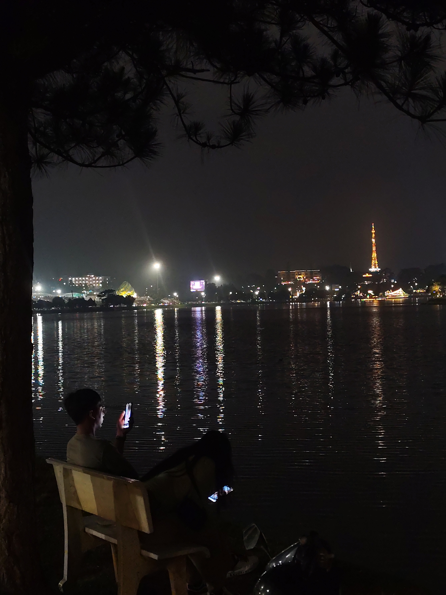 A dimly lit, wide-angle shot captures a nighttime scene at Ho Xuan Huong Lake in Da Lat, Vietnam. In the foreground, a person sits on a wooden bench under a tree, holding and looking at a mobile phone. Another person is seated next to them. A motorcycle is parked nearby. Across the lake, city lights reflect on the water's surface, with notable buildings and a tower illuminated in the distance.