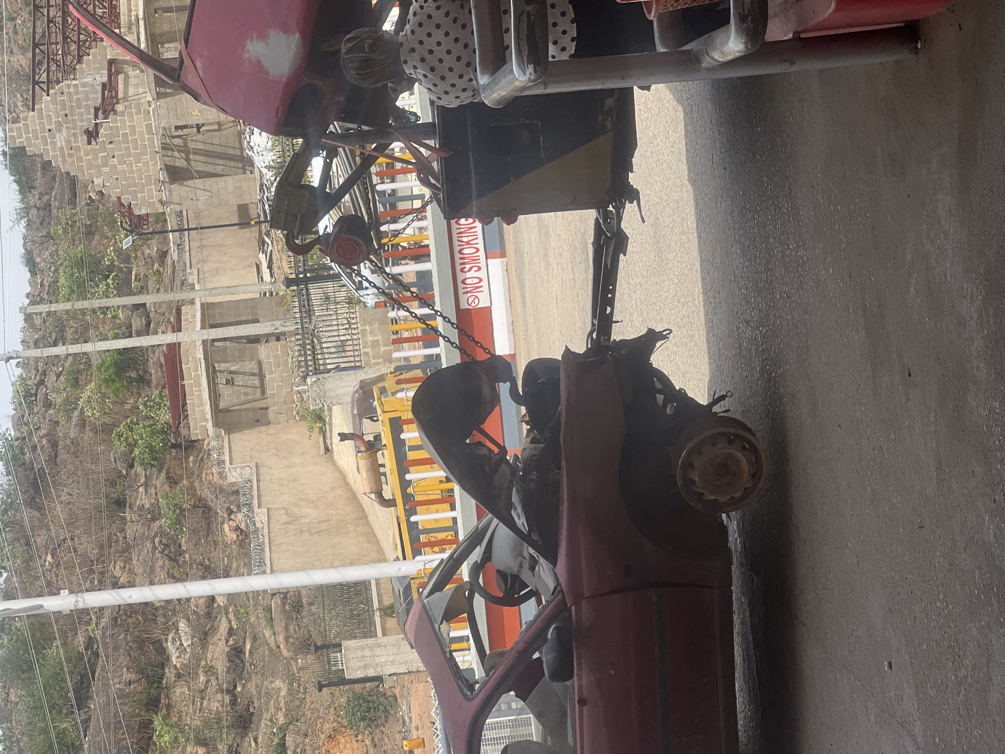The image shows a red, damaged car being lifted by a tow truck in what appears to be Jos, Nigeria. The car's front end is heavily damaged, with missing body panels and a bent frame. The tow truck is partially visible, with chains attached to the car's frame. In the background, there is a hillside with vegetation, a partly constructed building, and a "NO SMOKING" sign. The scene appears to be located on a paved road.