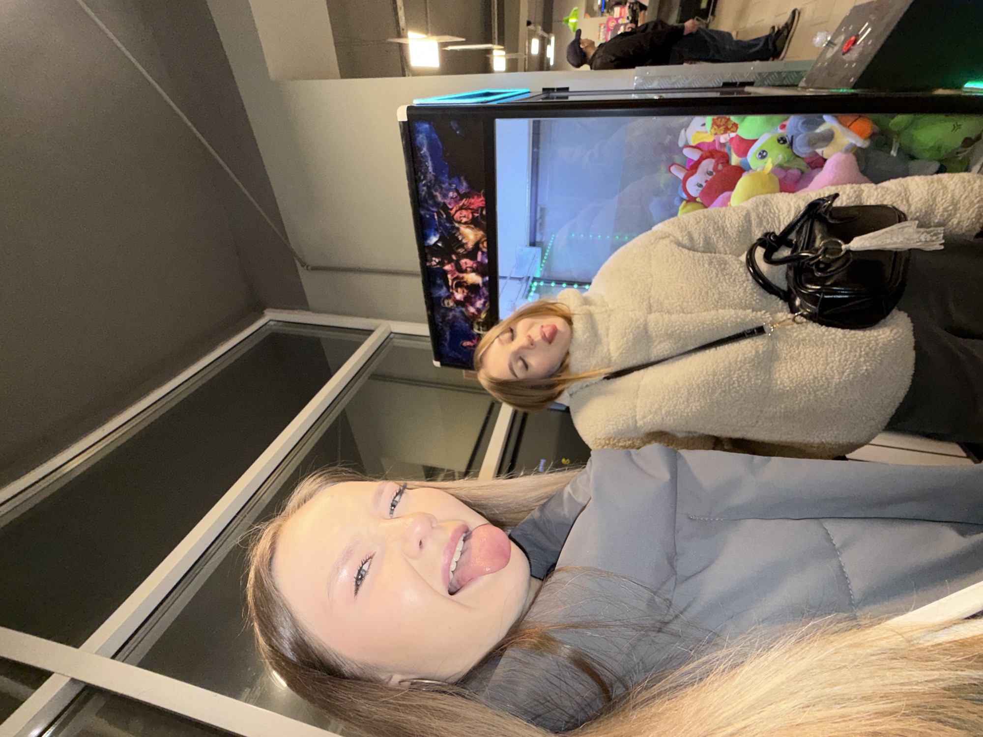 This image, taken in Chaikovsky, Russia, features two young women in a playful indoor setting, likely an arcade or entertainment venue. In the foreground, a woman with long light-brown hair, wearing a grey puffer jacket, looks towards the camera with her tongue playfully extended, displaying a happy and candid expression.
Behind her, another young woman is visible, wearing a cream-colored, fluffy shearling jacket and also has her tongue slightly out. She carries a black shoulder bag with a white tassel. Both women appear to be posing for a selfie or casual photo.
In the background, a large claw machine filled with various colorful stuffed animals, including red, green, and pastel-colored plush toys, dominates the scene. The side of the machine features abstract, colorful graphics. Further behind the claw machine, a man in dark clothing and a cap is seen lying down on what appears to be the top surface of another arcade game or structure.
The setting is indoors, characterized by artificial lighting, suggesting it might be evening or night. The ceiling features visible light fixtures and architectural details. Large windows or glass partitions are present on the left side of the frame. The overall atmosphere is casual and lively. No distinct text is visible within the image.