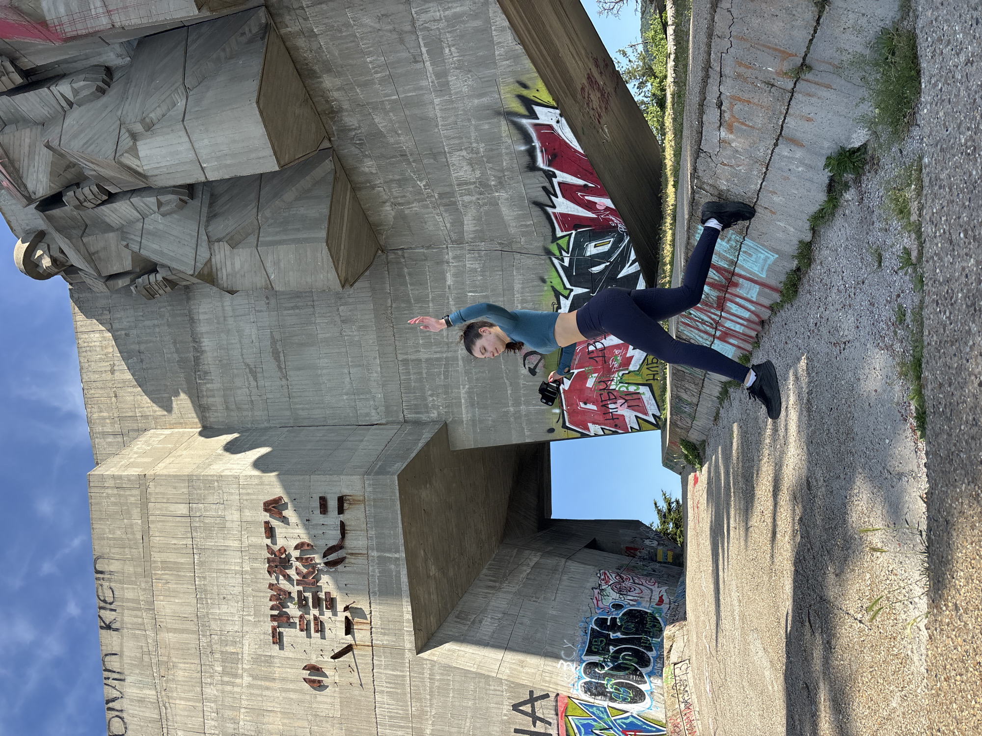 A woman wearing a blue long-sleeved shirt and dark leggings poses for a photo near the Monument of the Bulgarian-Soviet Friendship in Varna, Bulgaria. The woman, wearing black shoes, balances on the edge of a graffiti-covered concrete structure that forms part of the monument. The monument itself is a massive concrete edifice with abstract figures and lettering ("Lukea" is visible). The sky is visible in the background.